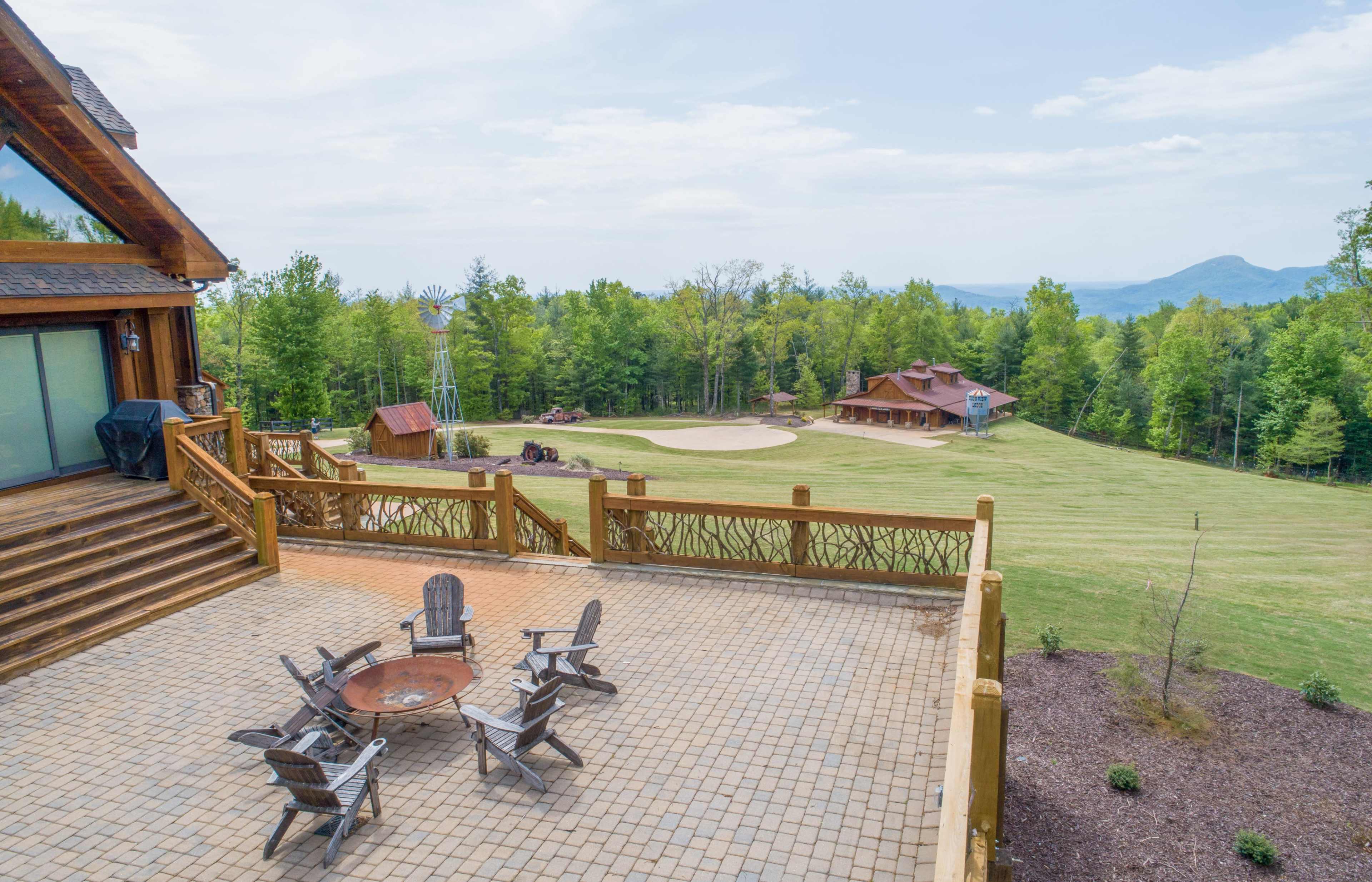 A spacious outdoor patio overlooks a large green lawn and a rustic log house surrounded by trees and mountains in the background.
