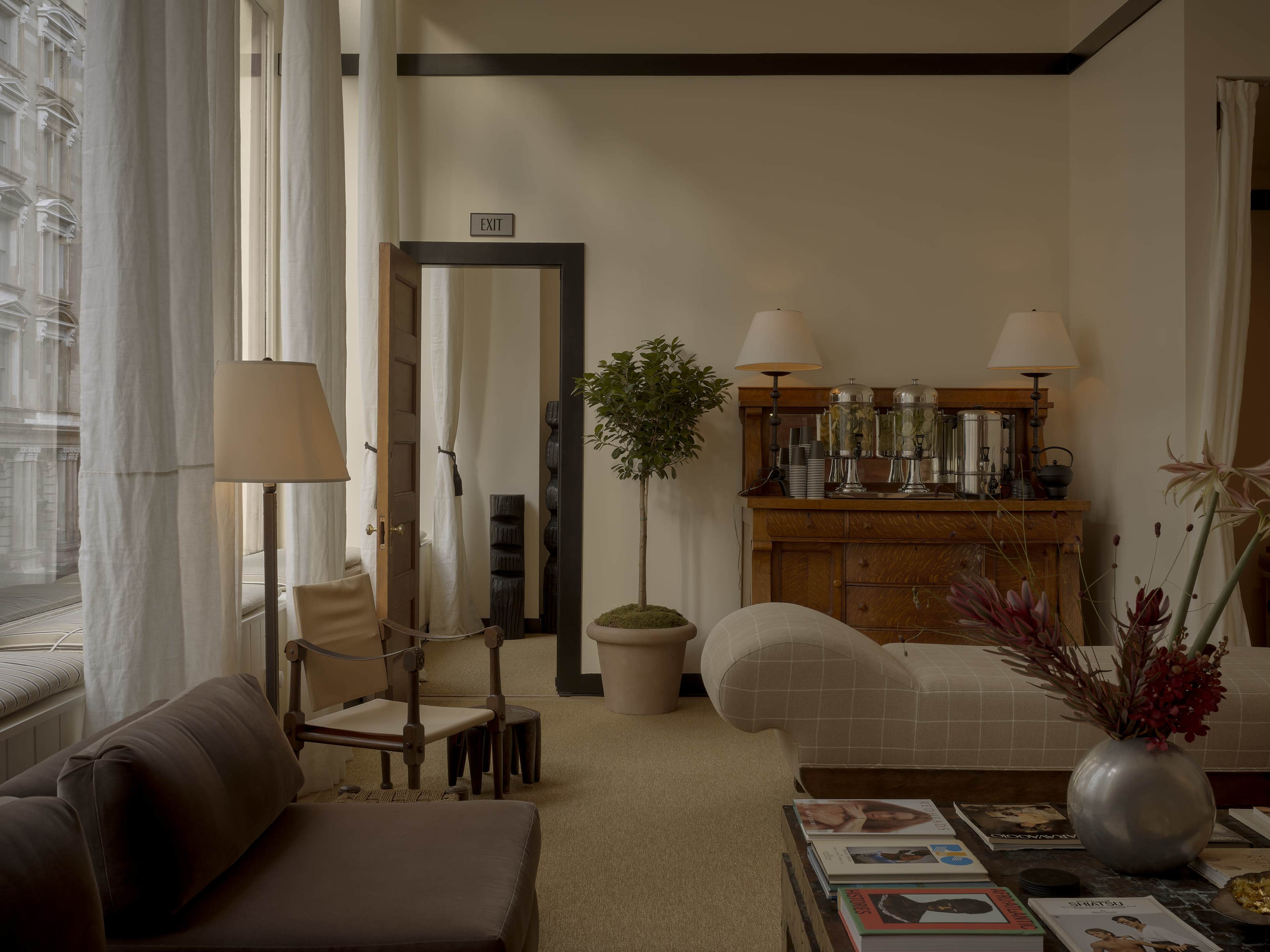 The image shows a stylish living room featuring a modern sofa, a wooden sideboard with a coffee setup, a plant, and large windows with light-colored curtains.