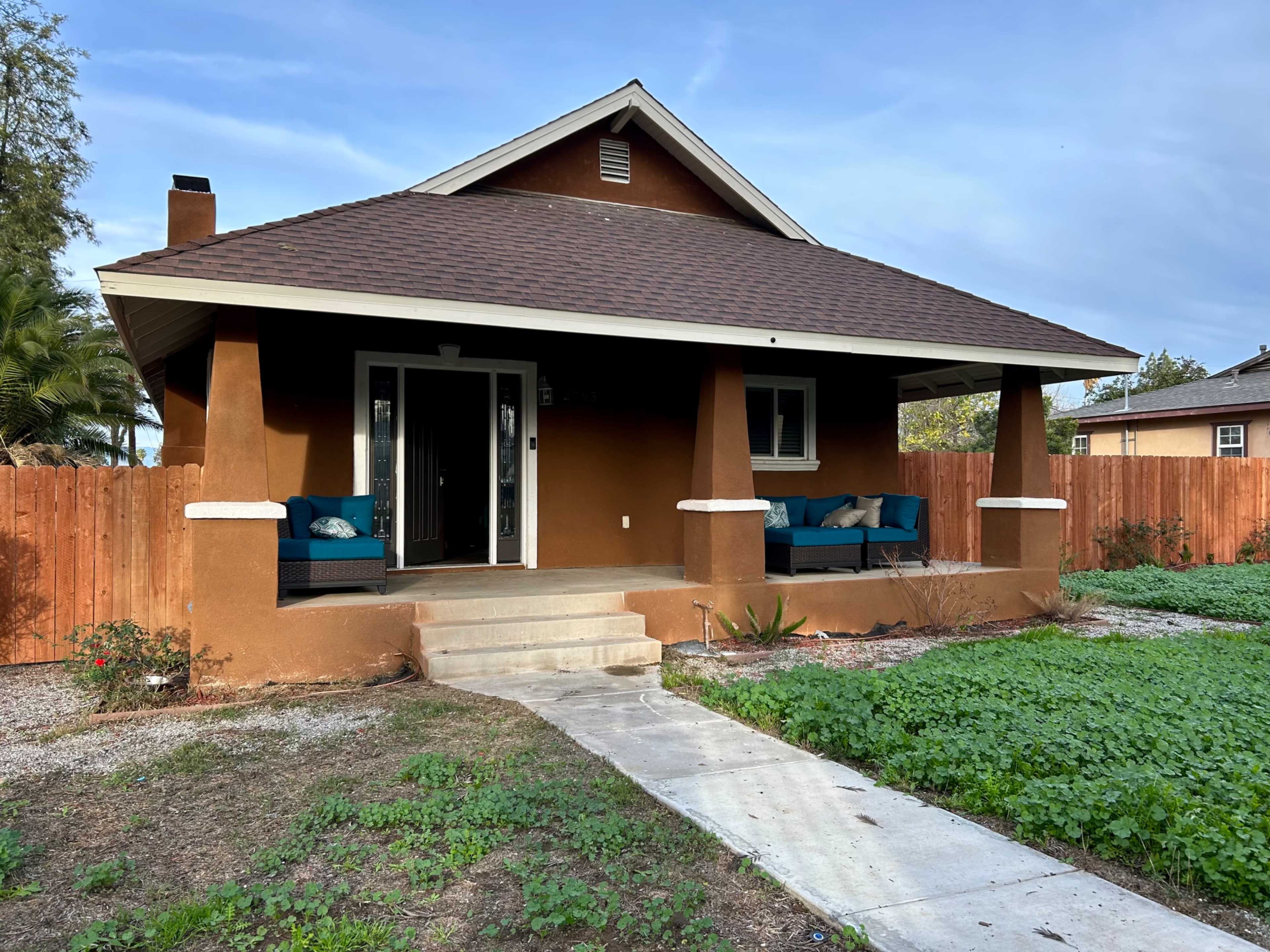 A brown bungalow with a front porch and two blue chairs sits in a yard with grass and gravel pathways.