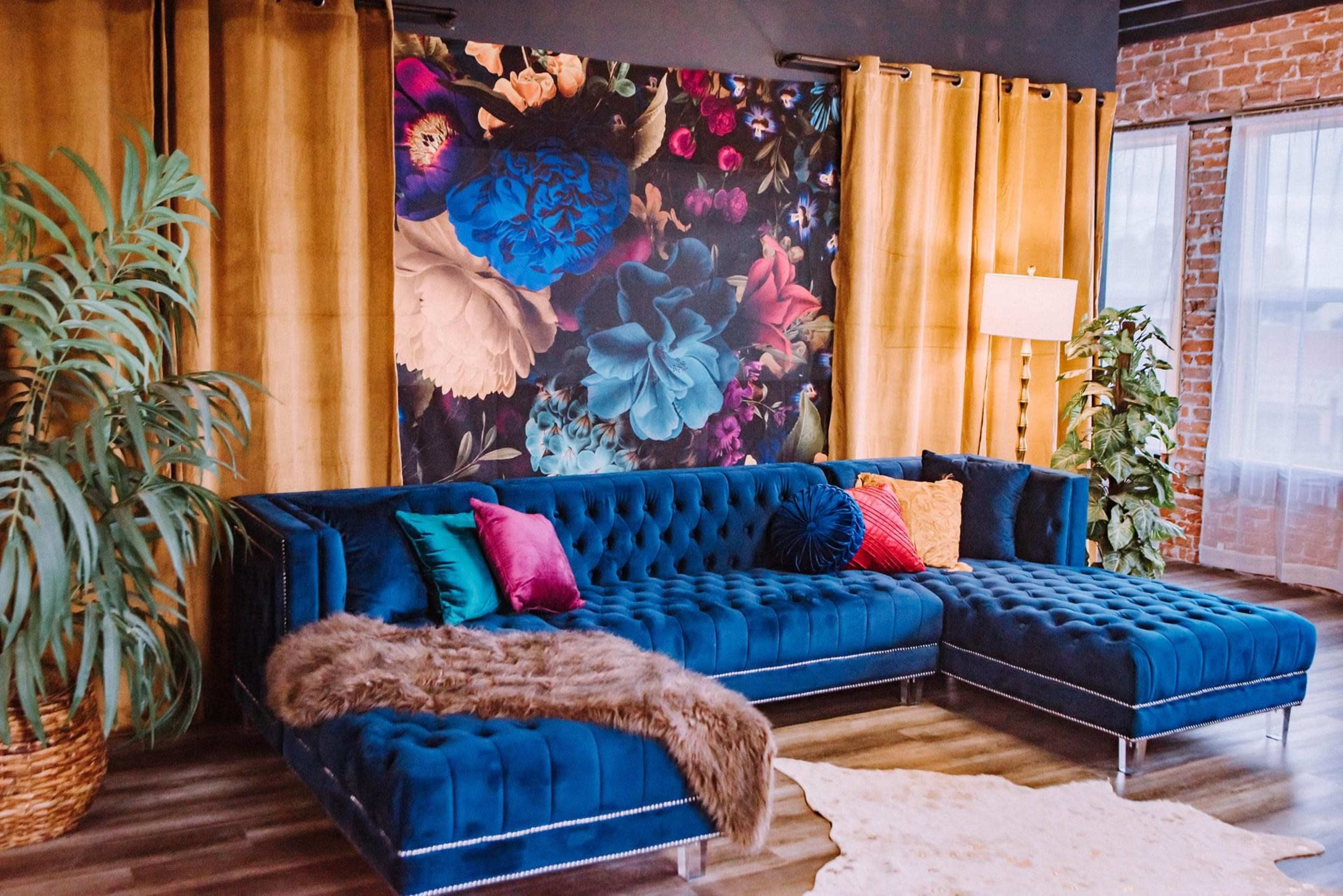 A modern living room with a blue velvet sectional sofa, colorful cushions, a decorative fur throw, and a large floral wall mural surrounded by yellow curtains.