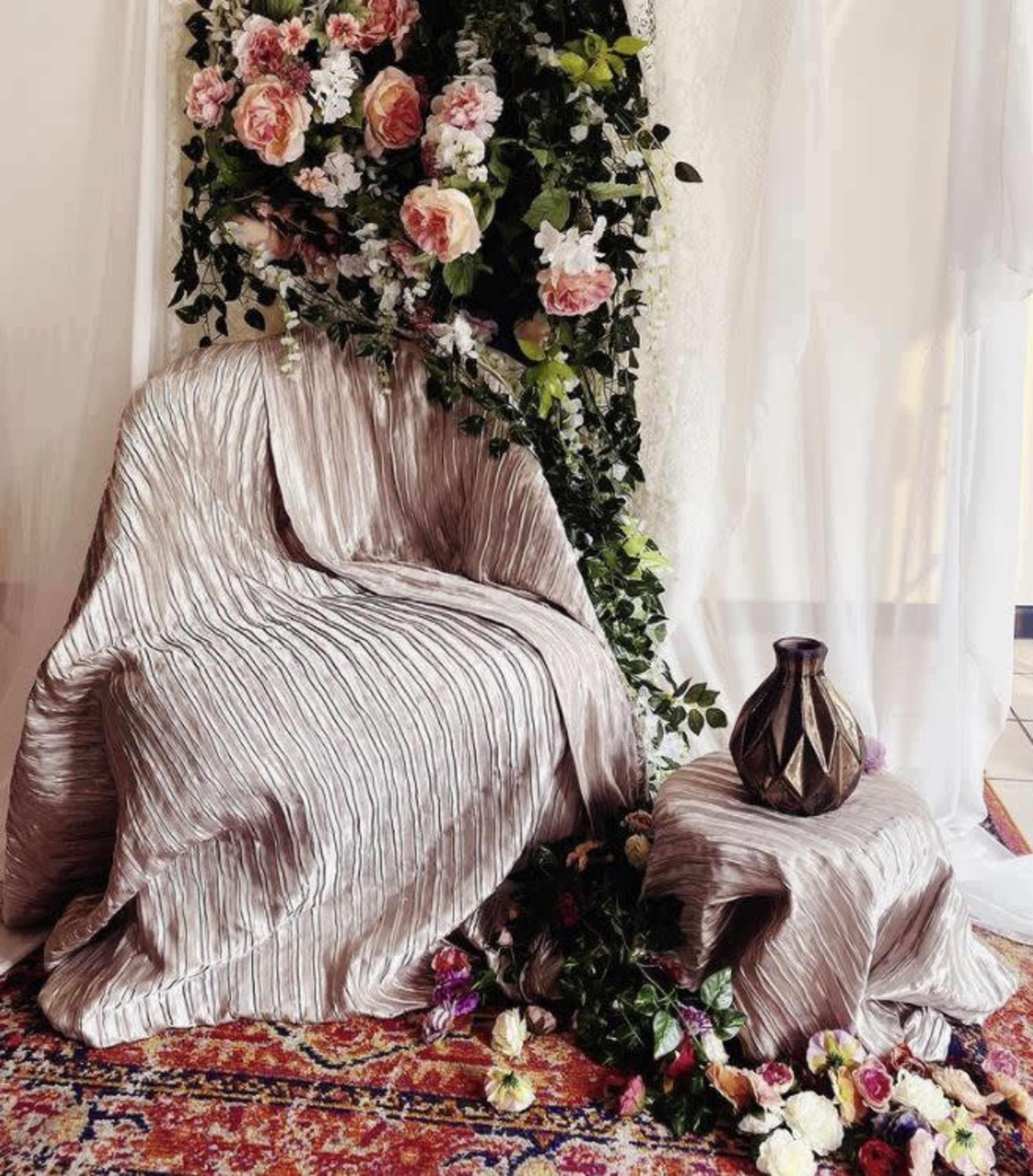 A fabric-draped chair and side table adorned with flowers and a decorative vase are positioned against a backdrop of white drapery.