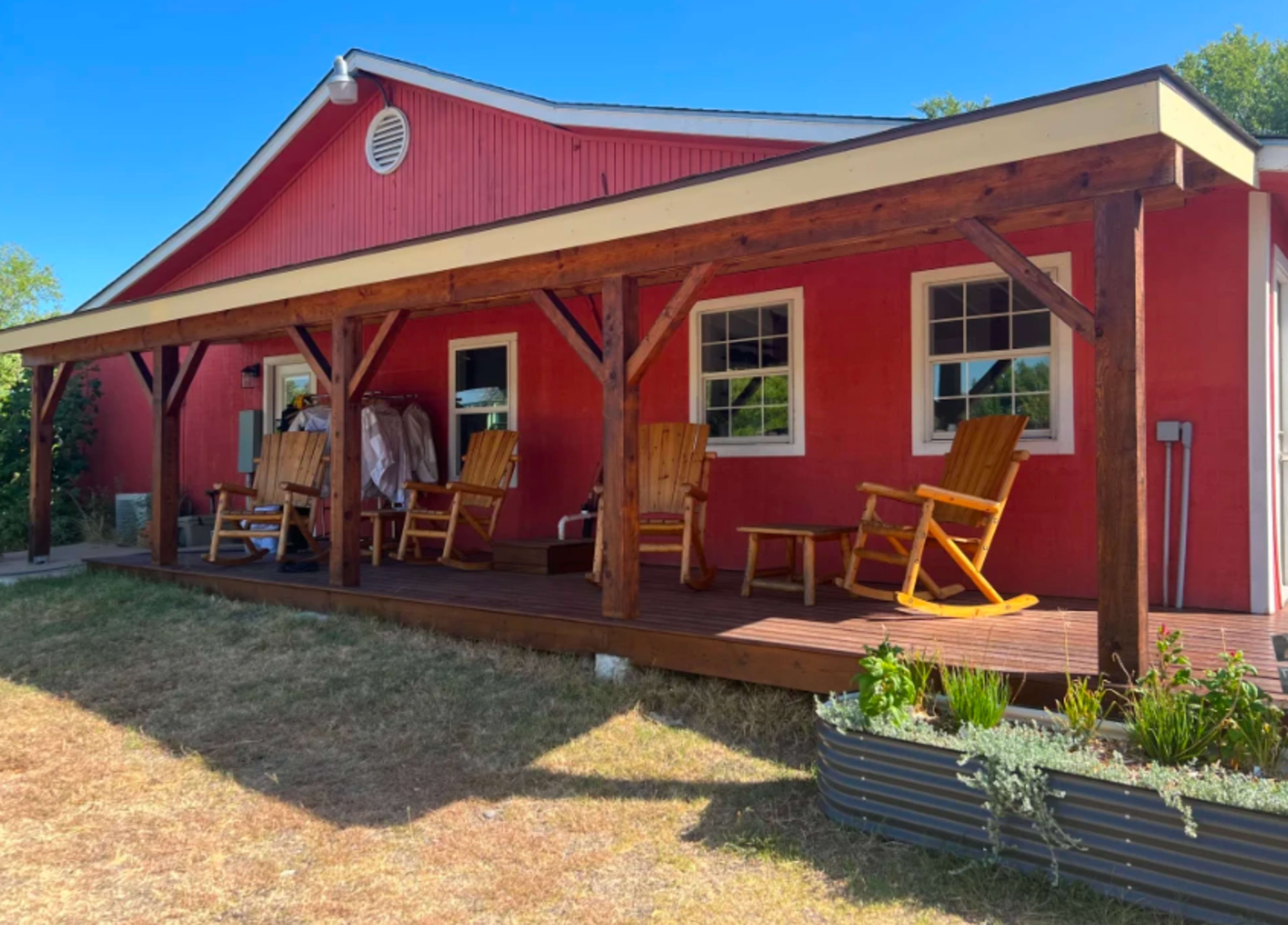 A red house with a wooden porch features several rocking chairs and plants along the side.