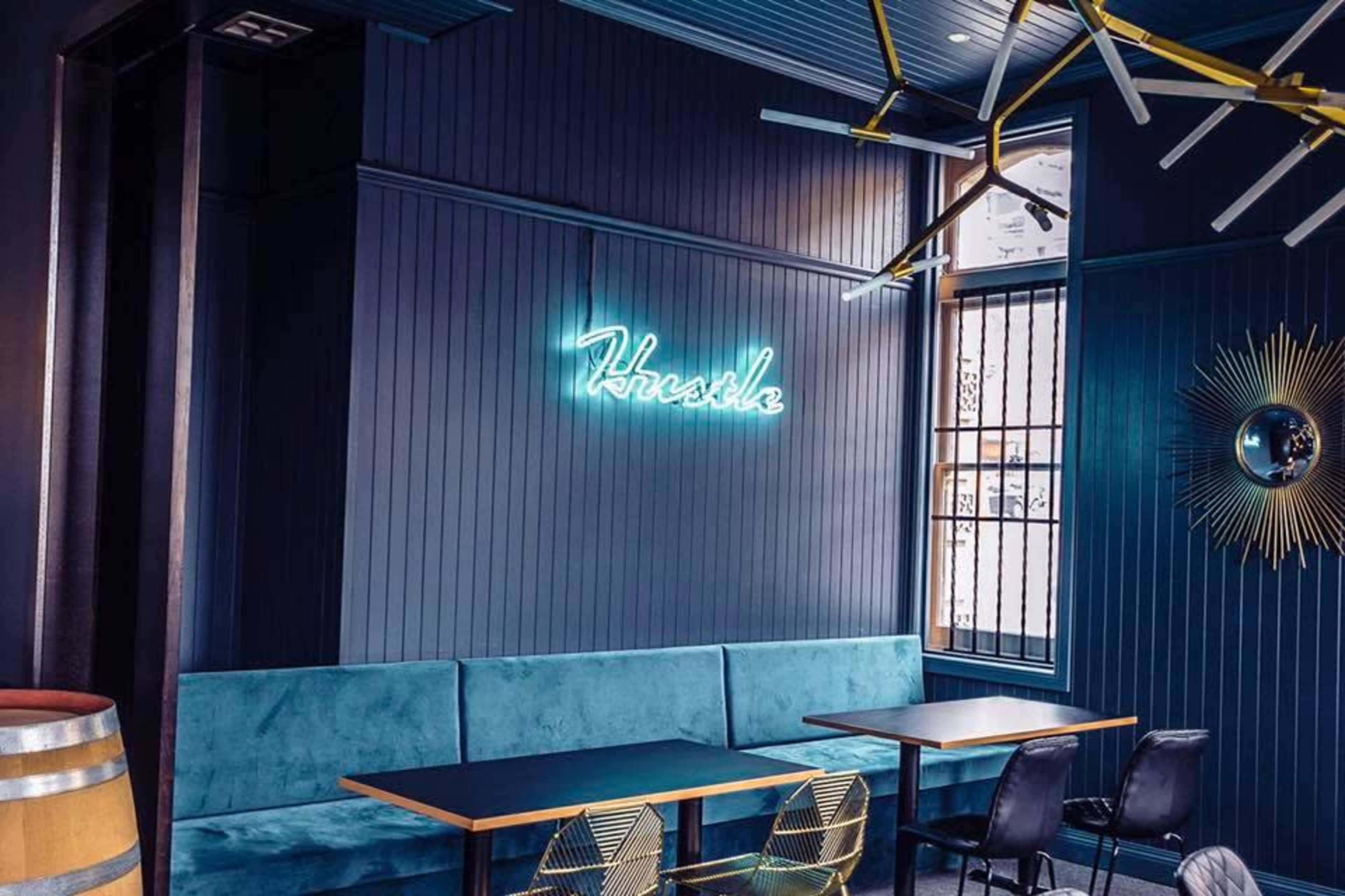 A modern cafe interior featuring a blue wall, a neon sign that says "Hustle," a long blue upholstered bench with wooden tables, and a mix of black and gold seating.