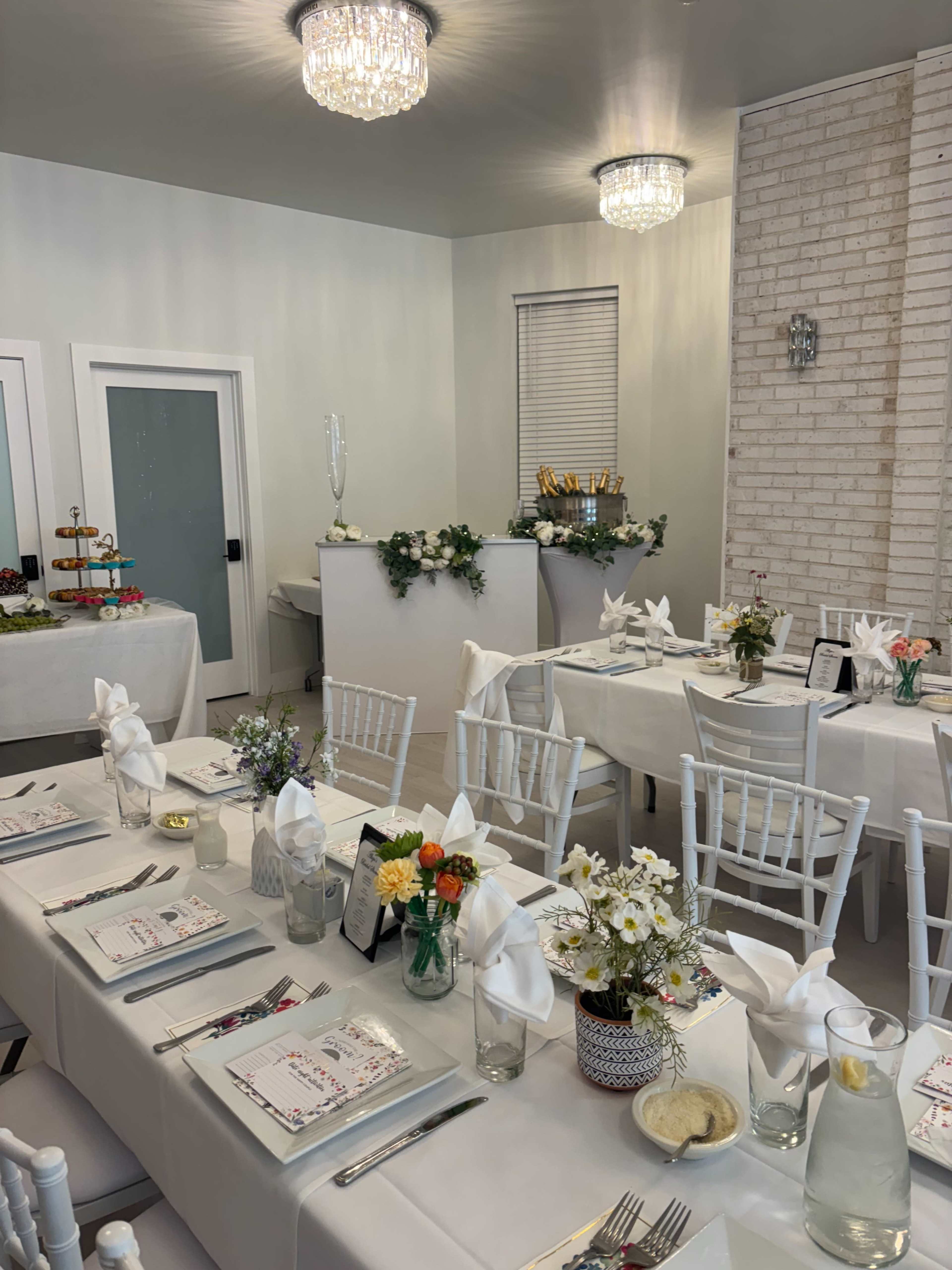 The image shows a neatly arranged dining area with white tables set with floral centerpieces, folded napkins, and place settings, complemented by decorative lighting and a buffet table in the background.