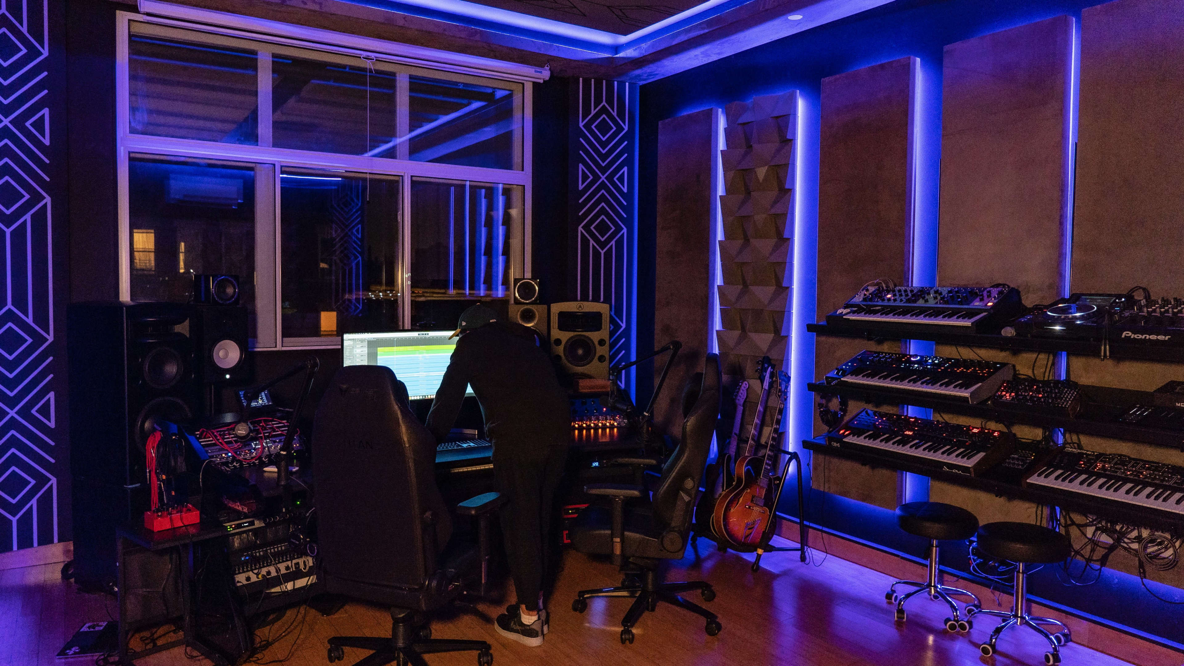 A person is working at a music production workstation in a studio filled with various musical equipment and illuminated by blue lights.