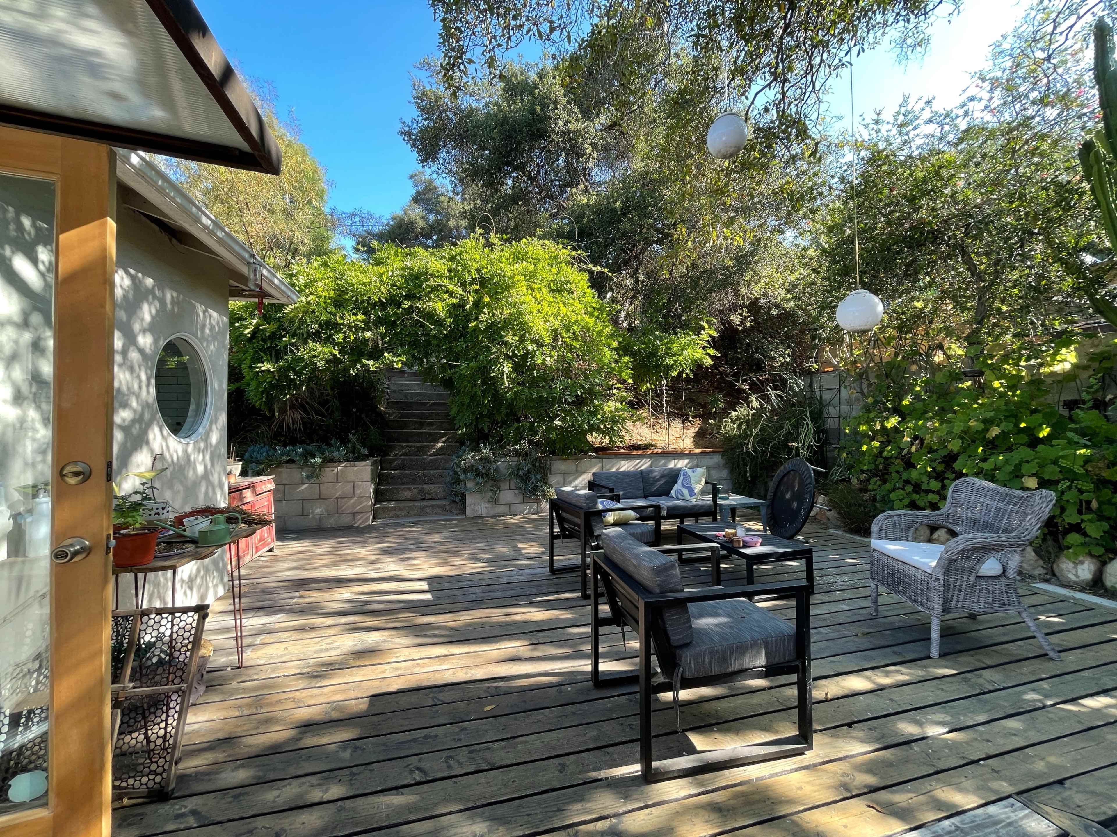 A wooden deck with outdoor furniture, surrounded by lush greenery and stairs leading to a garden area.