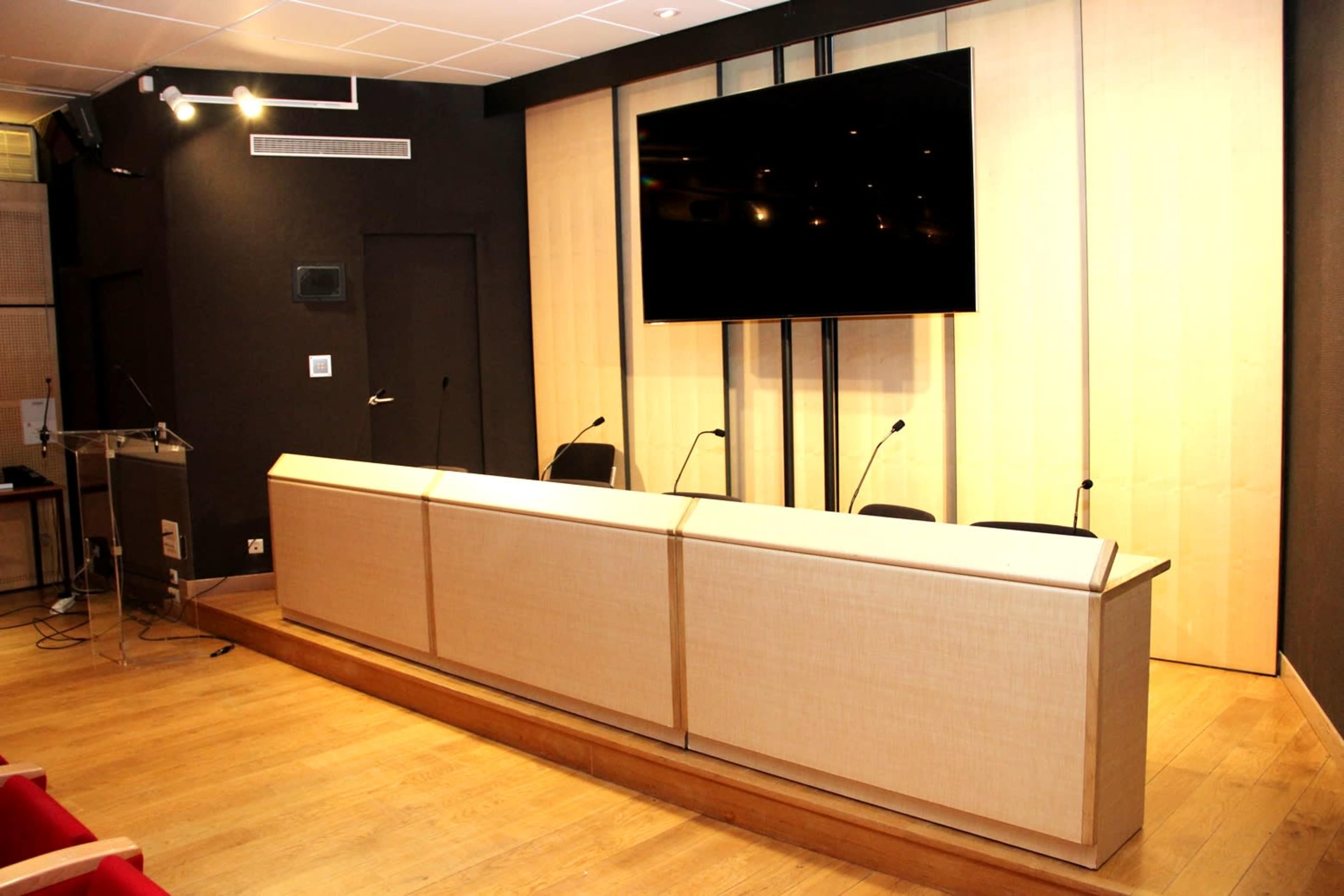 A conference room features a long table with multiple microphones, a large screen on the back wall, and several chairs arranged for a meeting.