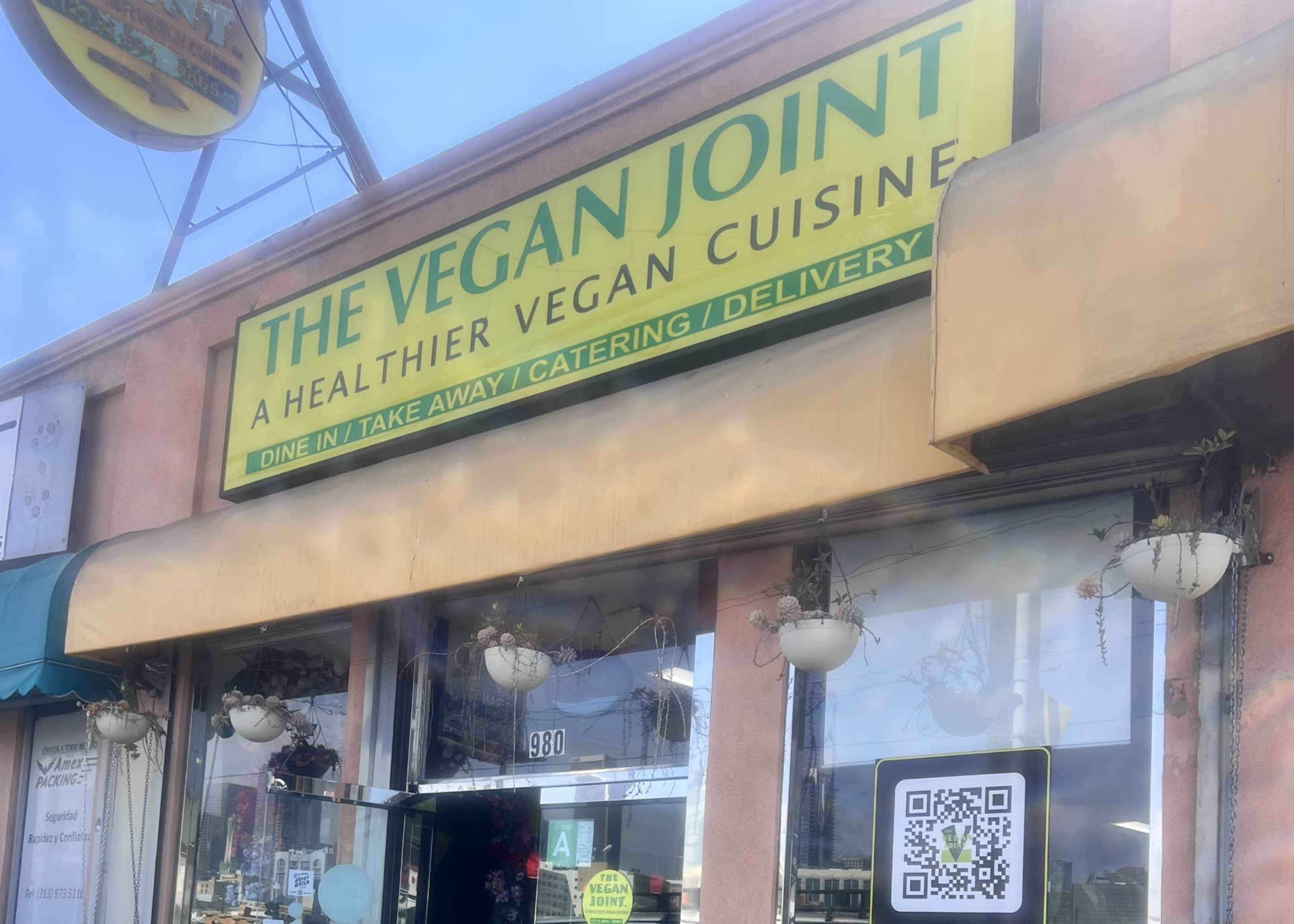 The image shows the exterior of a restaurant called "The Vegan Joint," featuring a green sign that emphasizes its vegan cuisine and services like dine-in, take away, catering, and delivery.