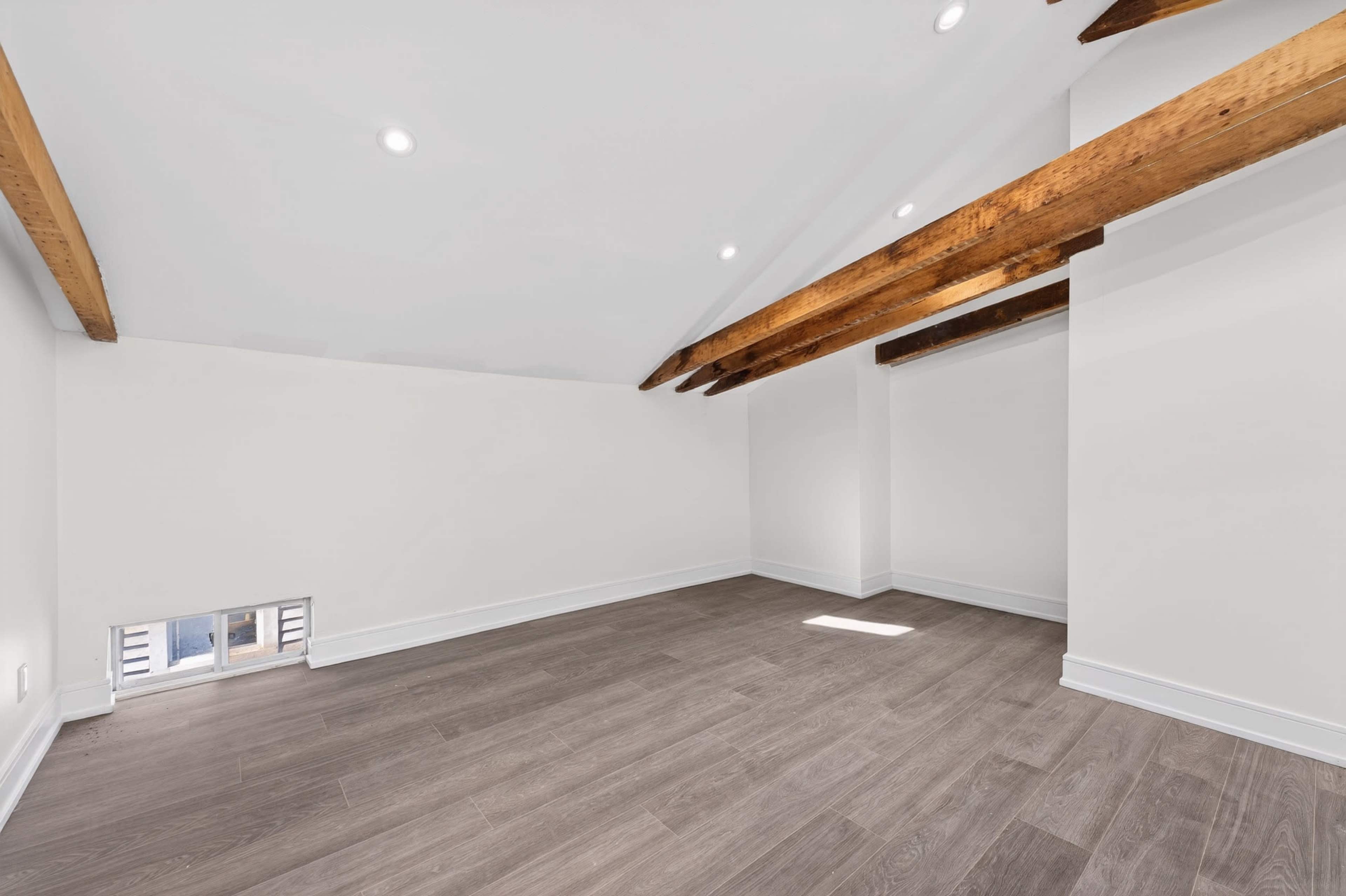 The image depicts an empty room with wooden beams on the ceiling, pale walls, and laminate flooring.