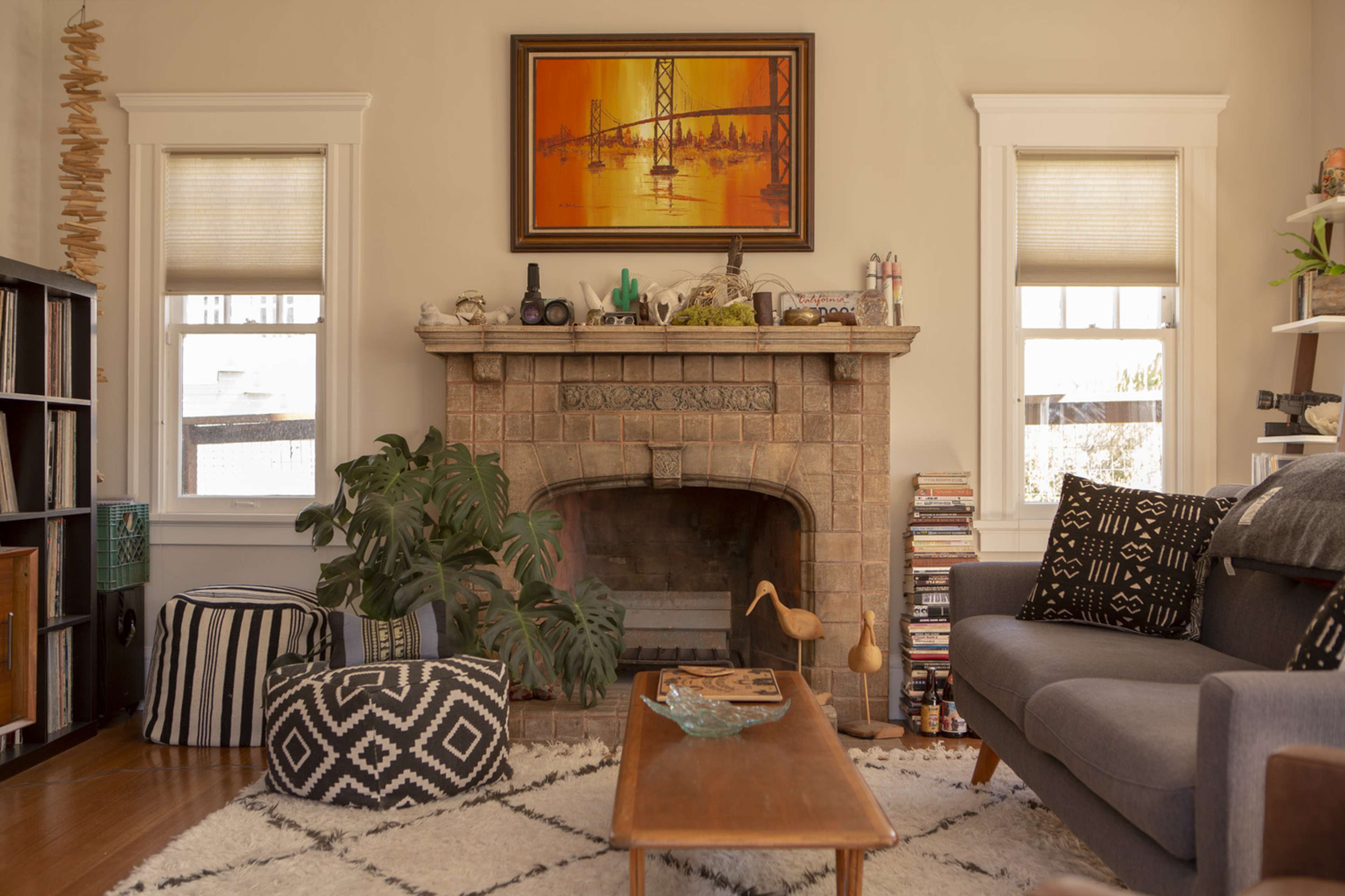 The living room features a stone fireplace with a painting above it, a gray sofa, patterned ottomans, and a small coffee table surrounded by plants and shelves filled with books and decor items.