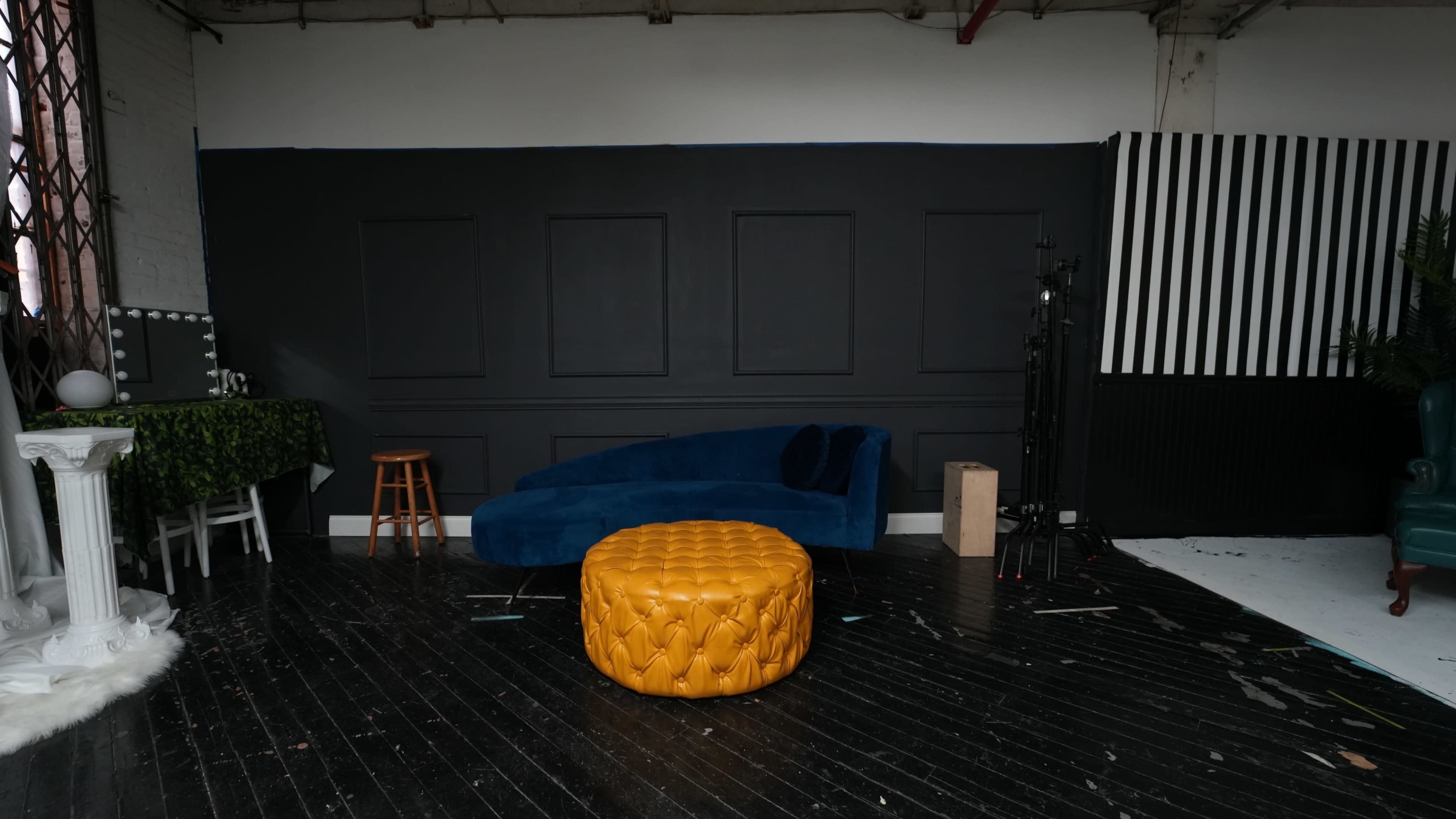 A spacious studio with a dark wall, a blue couch, a yellow ottoman, and various backdrops and props arranged around the room.