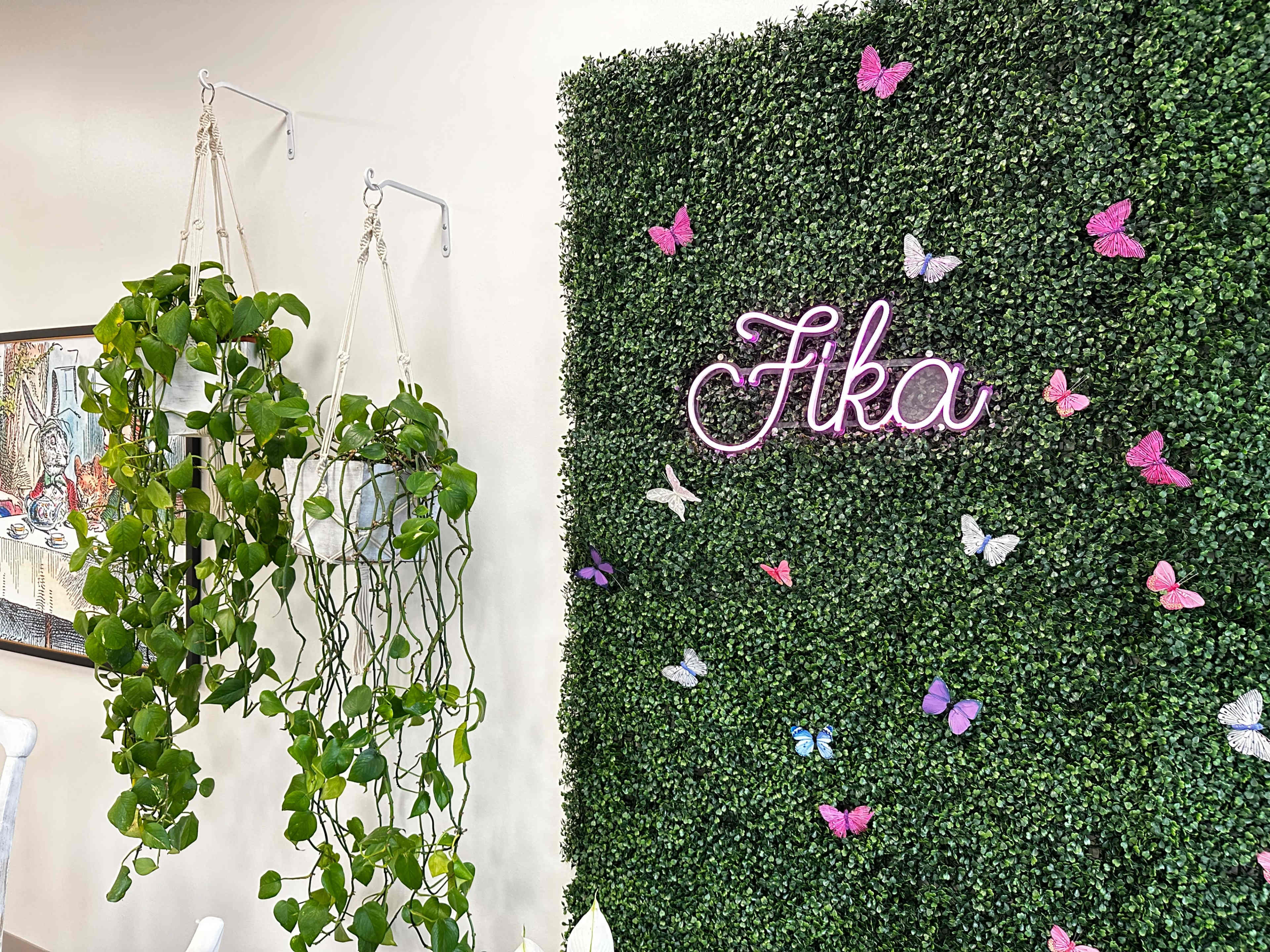 A decorative wall covered with artificial greenery, featuring a neon sign that reads "Fika" and several colorful butterfly decorations, alongside hanging plants in macramé holders.