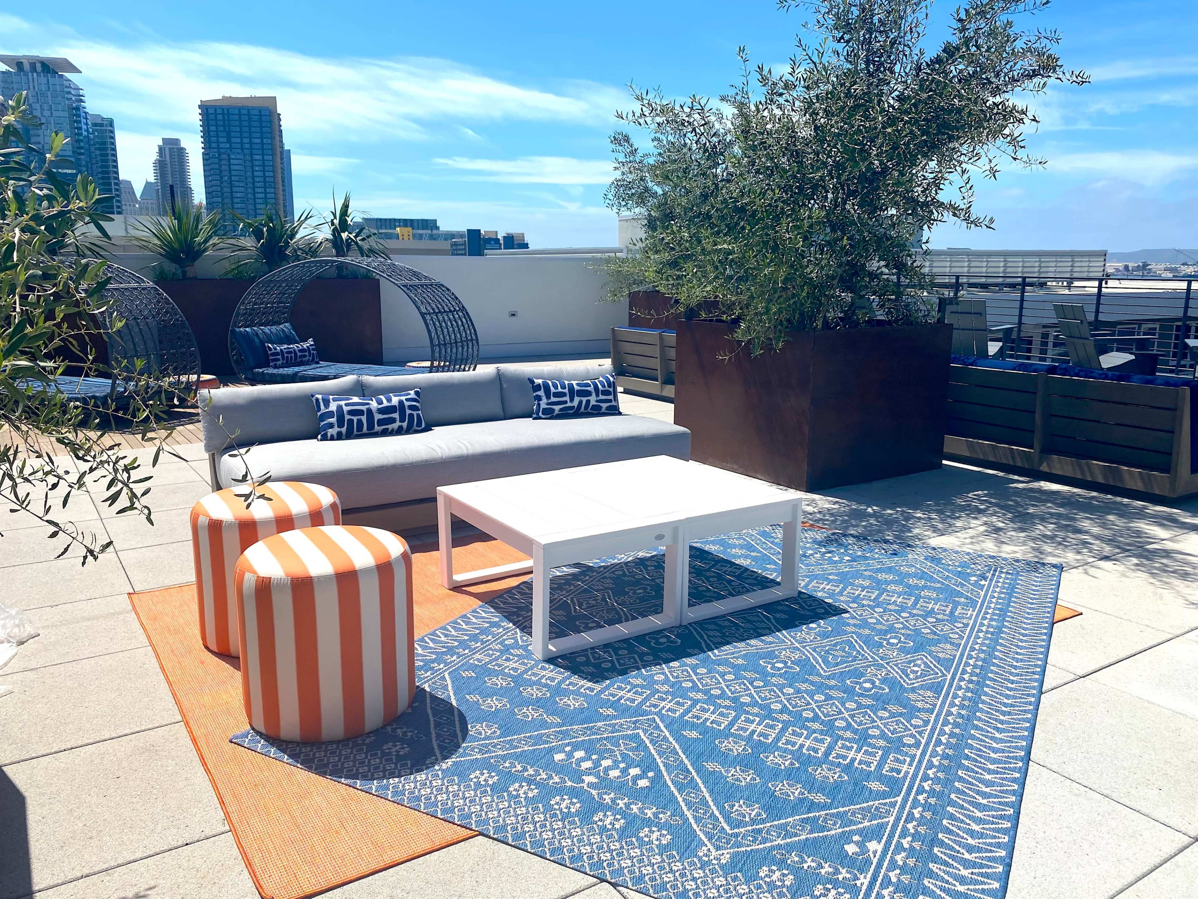 The image shows a rooftop seating area featuring a couch, patterned stools, a coffee table, and decorative plants under a clear sky.