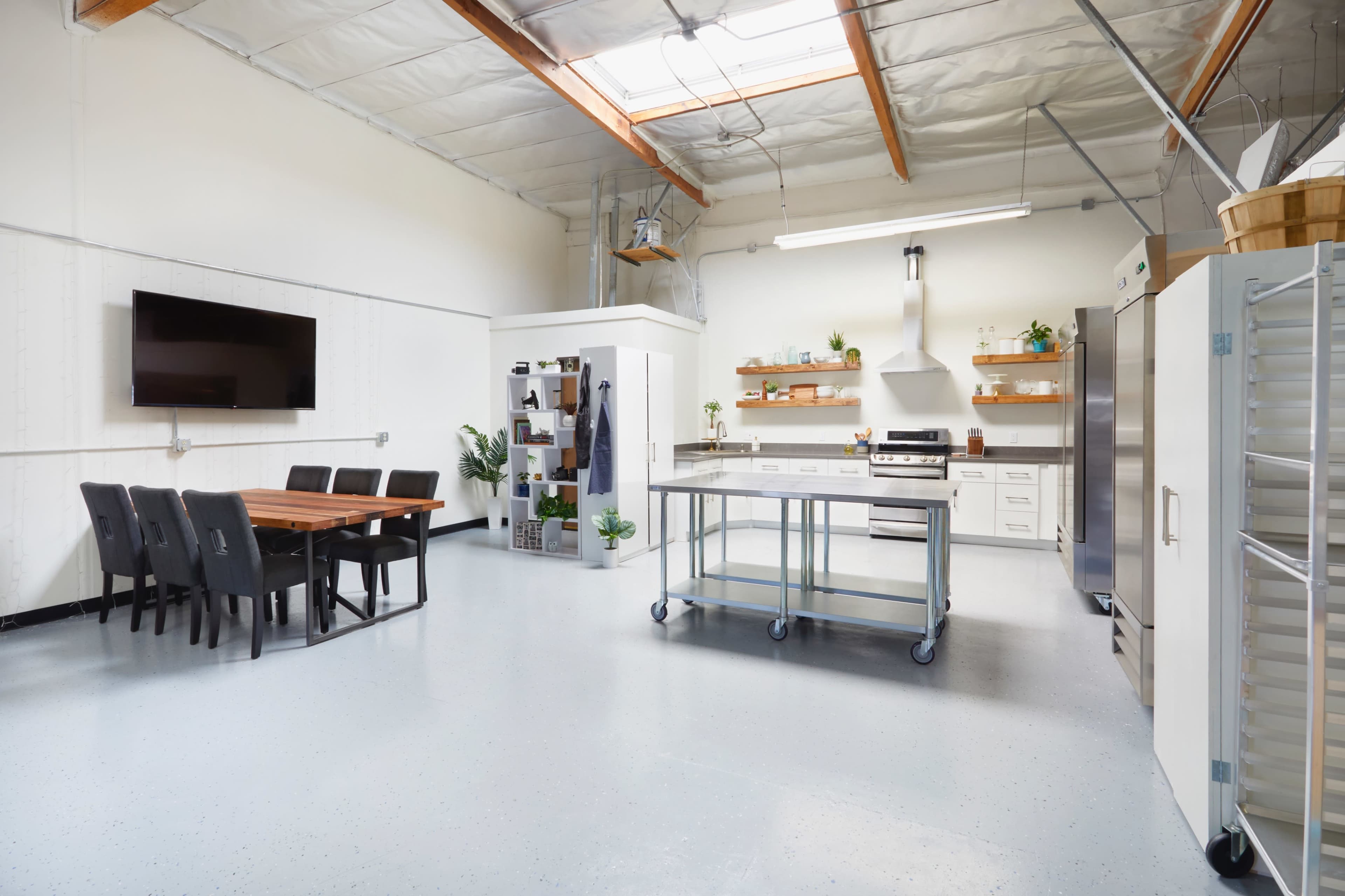 The image shows a spacious, modern kitchen with a dining area, featuring a large table, stainless steel appliances, and plants on open shelves.