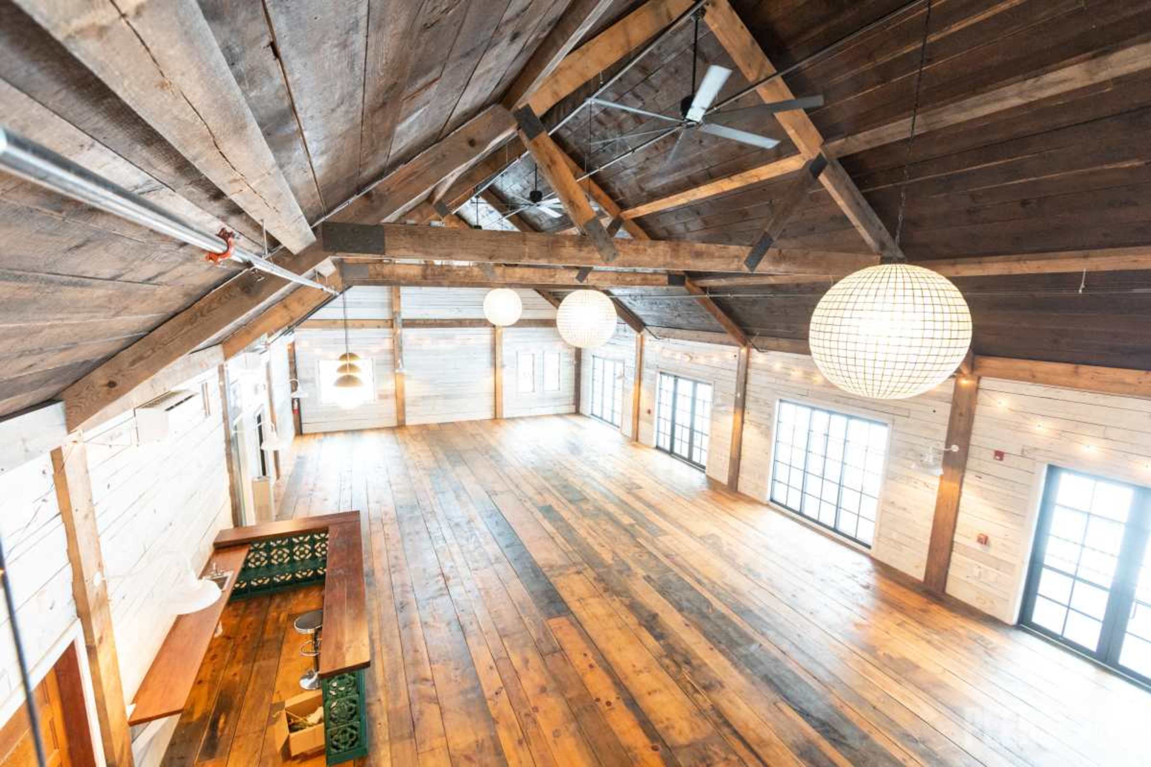 The image shows a spacious, wooden interior of a building with high ceilings, large windows, and overhead decorative lights.
