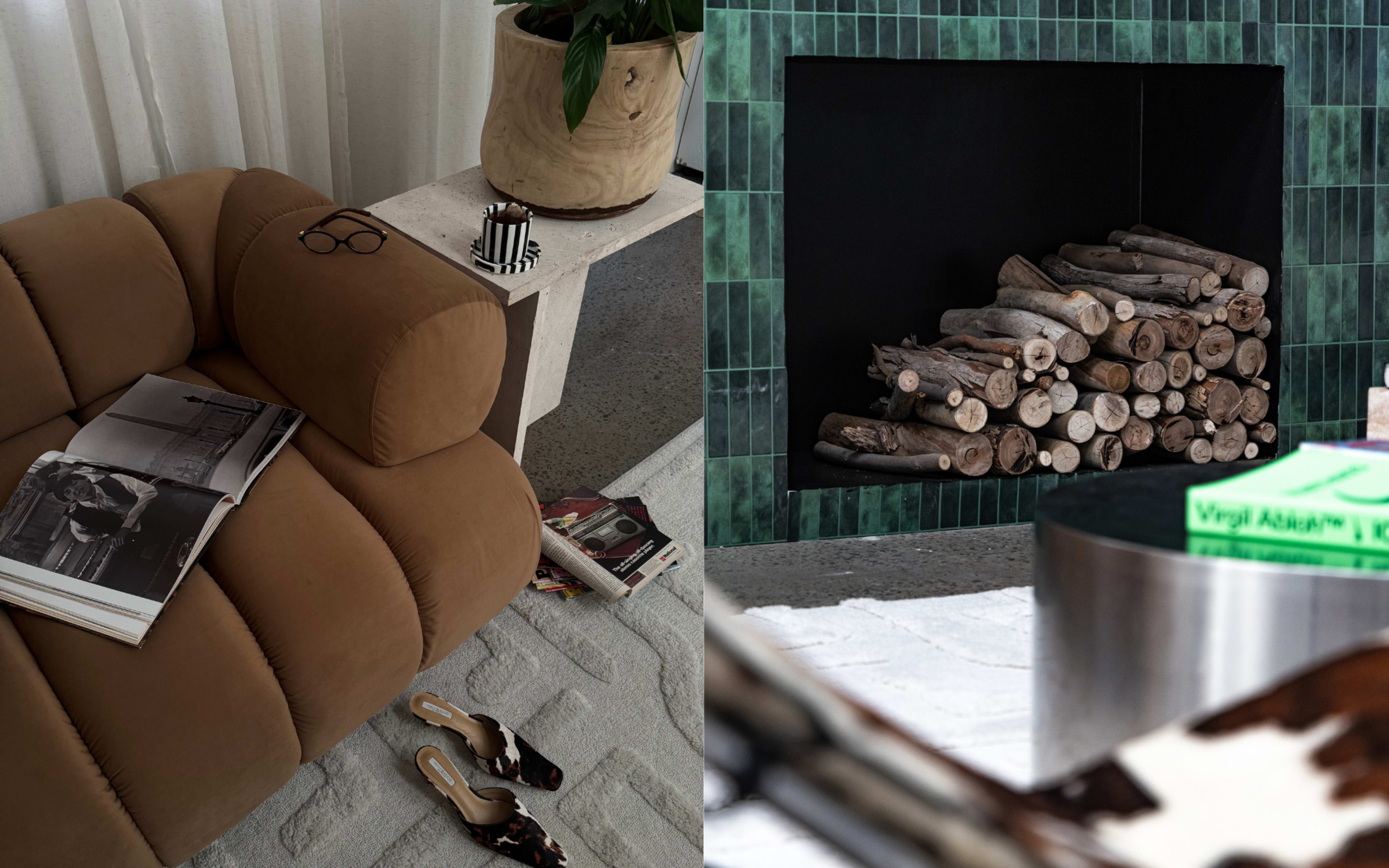 The image shows a cozy living room scene featuring a modern, tan sofa with magazines and a pair of glasses on it, alongside a neatly stacked pile of firewood in a black fireplace surrounded by green tiled walls.