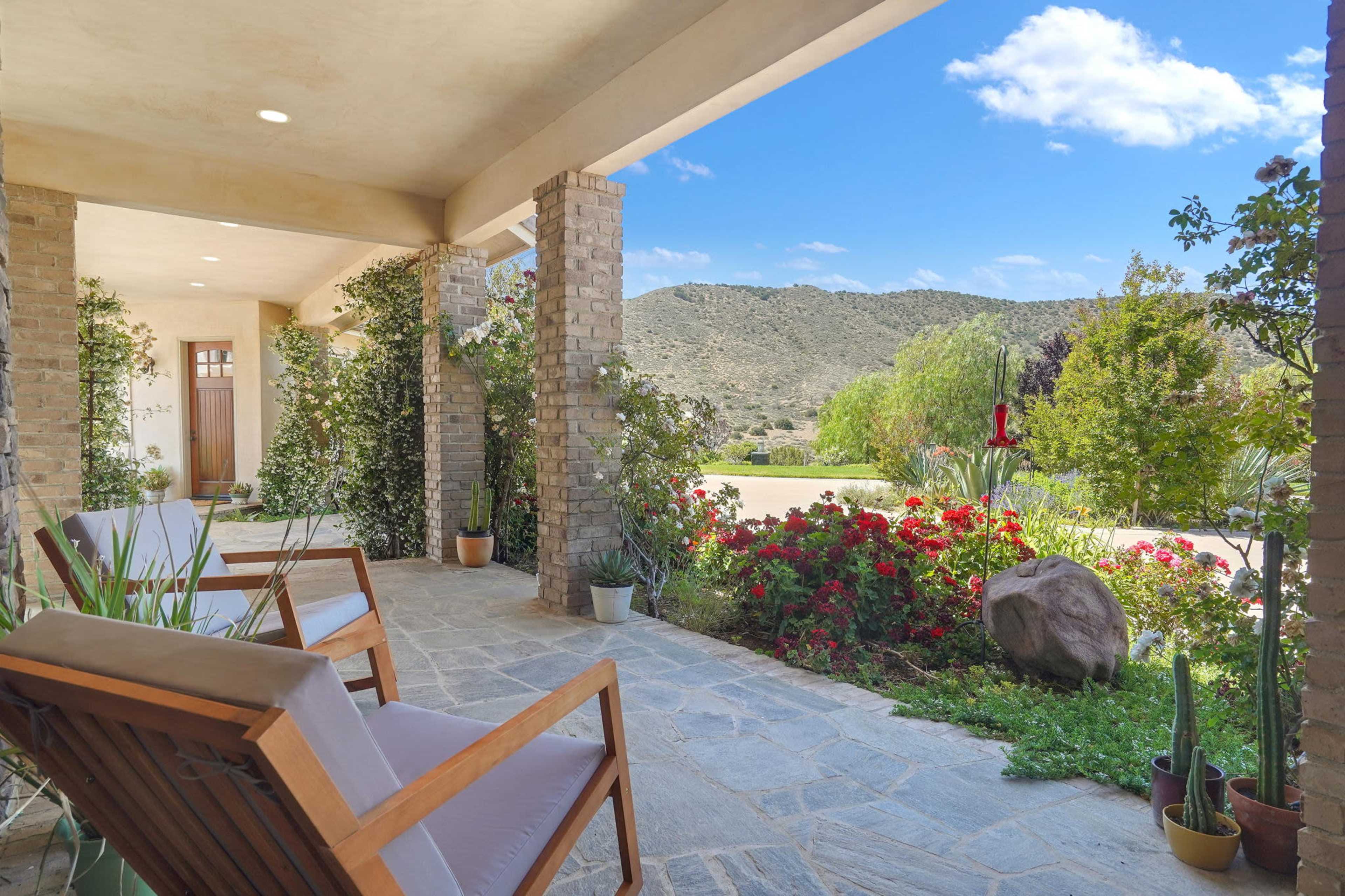 The image shows a covered porch with wooden chairs overlooking a garden filled with flowers and a mountainous landscape in the background.