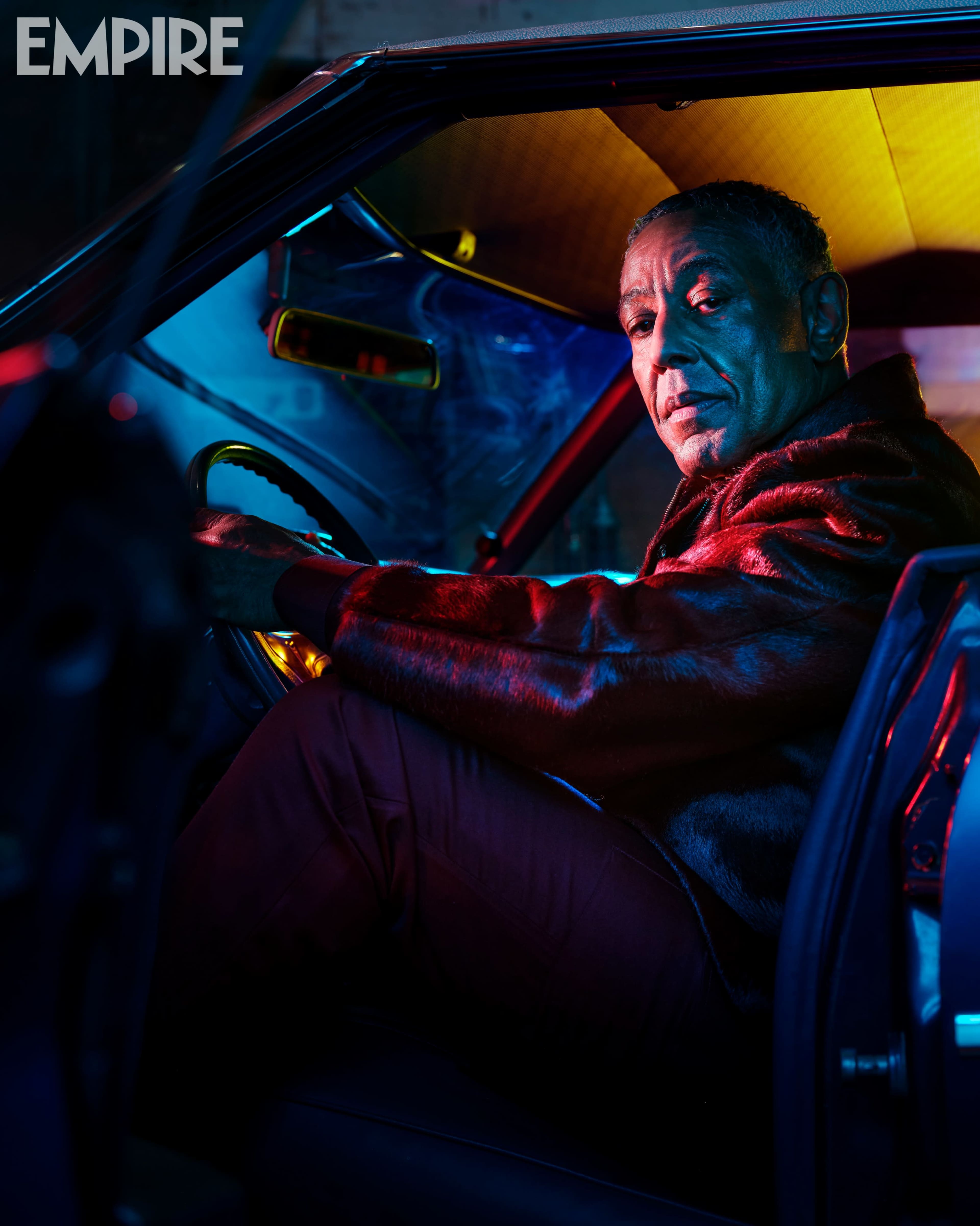 A person sits inside a vintage car, illuminated by colorful lighting and looking directly at the camera.