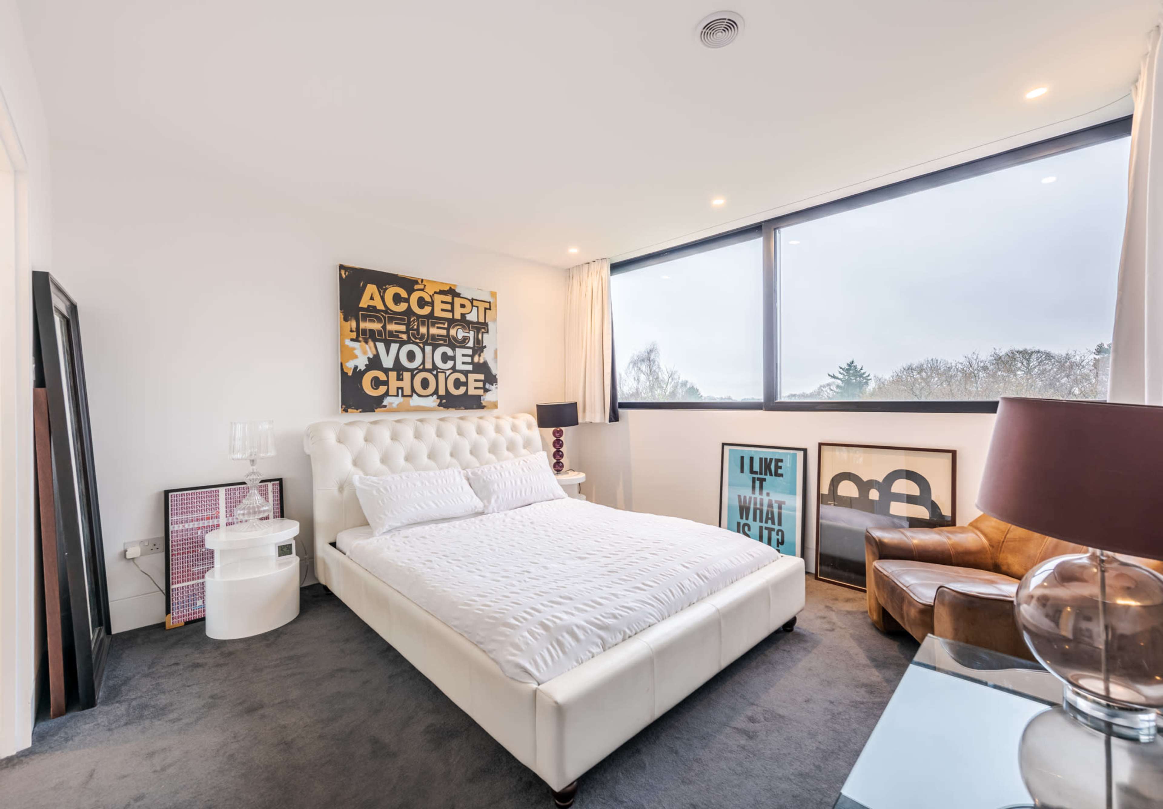 A modern bedroom features a large white bed, a brown leather chair, and artwork on the walls, illuminated by natural light from a large window.