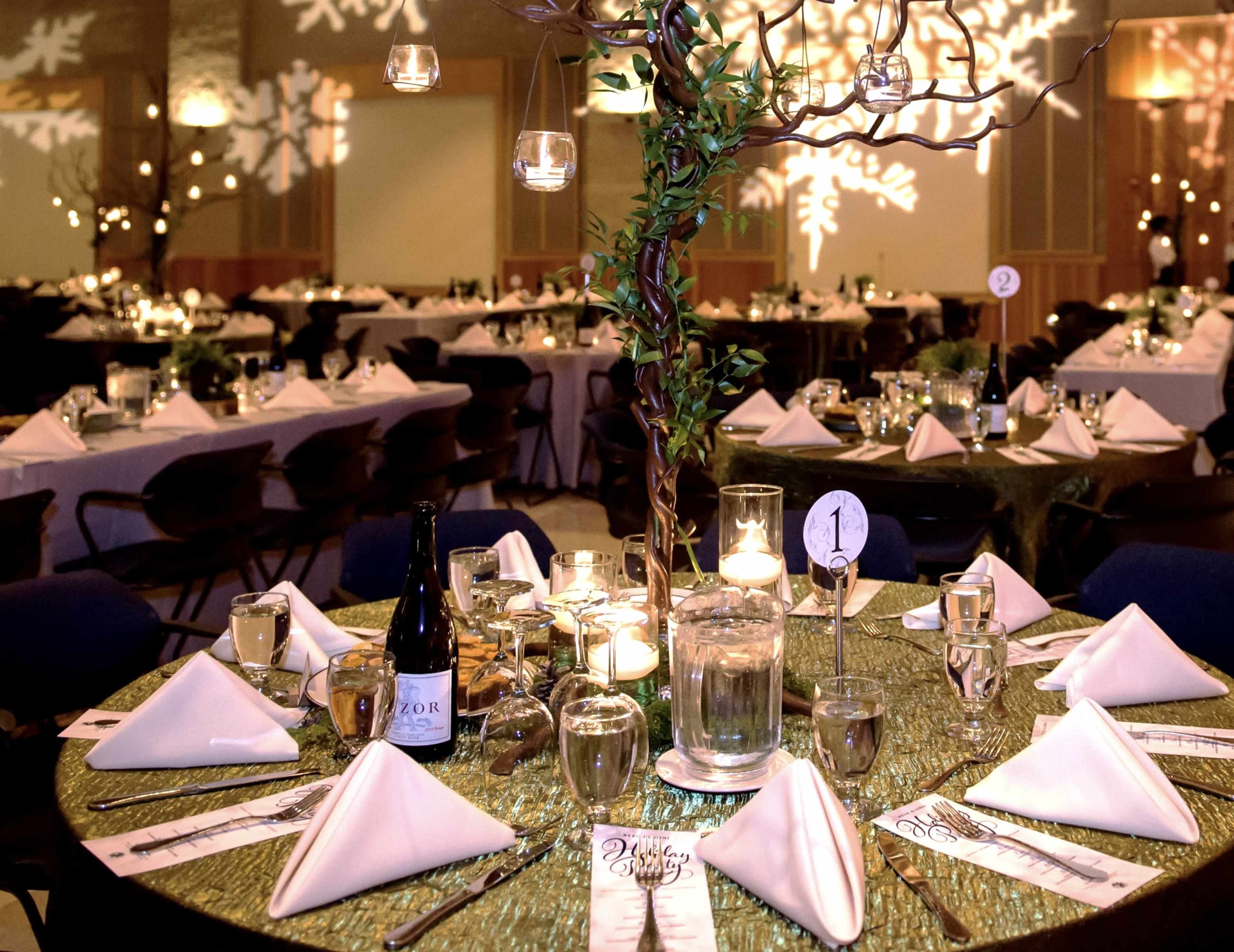The image shows a beautifully arranged banquet hall with round tables set with elegant dinnerware, napkins, and a centerpiece featuring a decorative tree adorned with hanging candles.