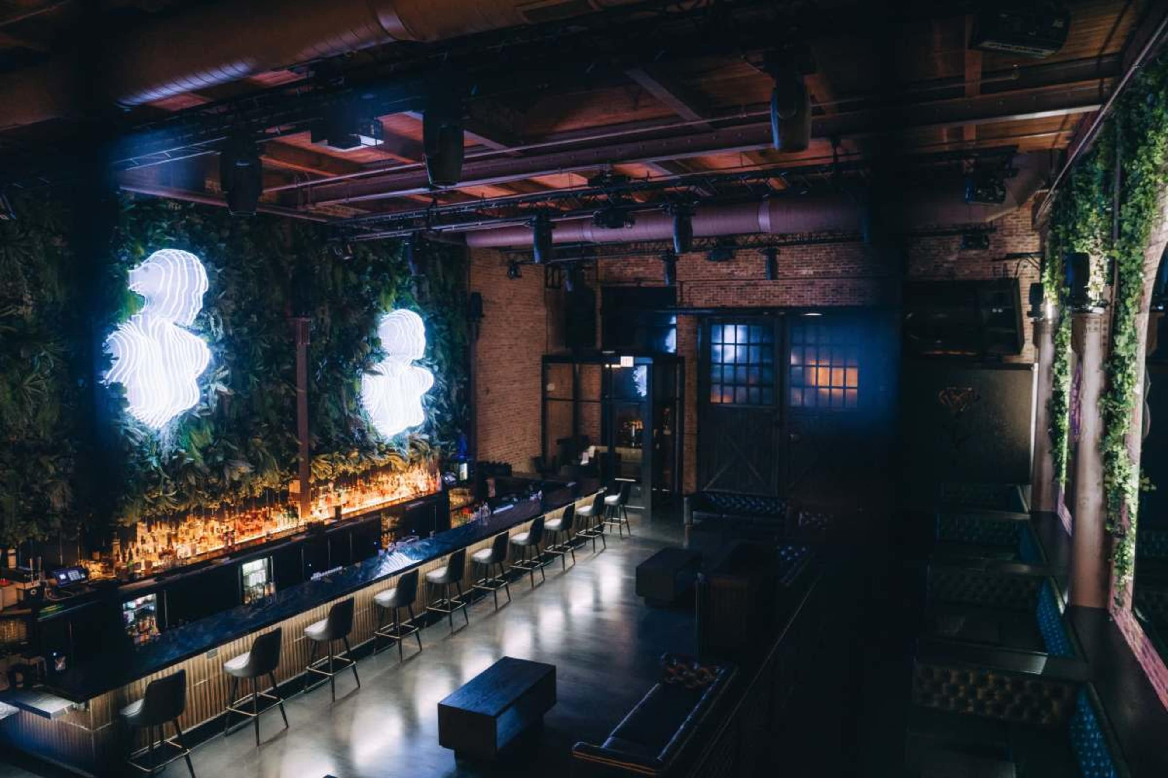The image shows a modern bar interior featuring a well-lit counter, lush green wall decor, and seating arrangements in a spacious setting.