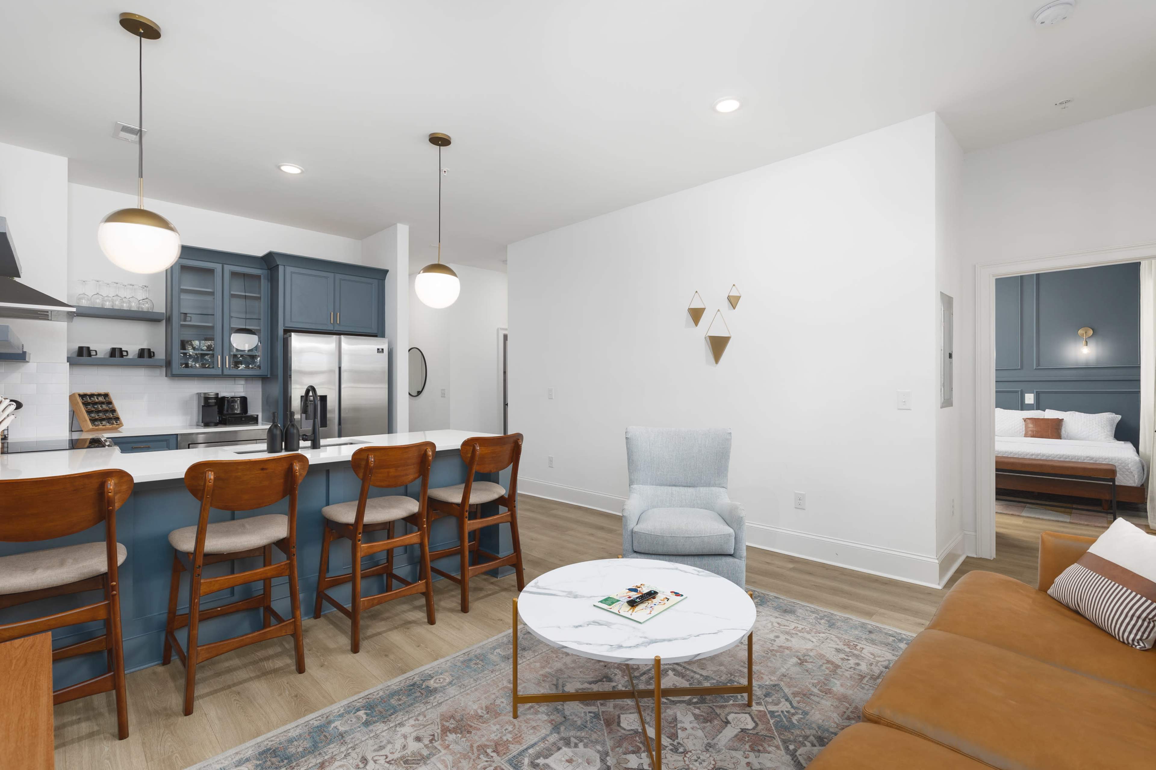 The image shows a modern living space featuring a kitchen island with barstools, a beige sofa, a marble coffee table, and a visible bedroom through an open door.