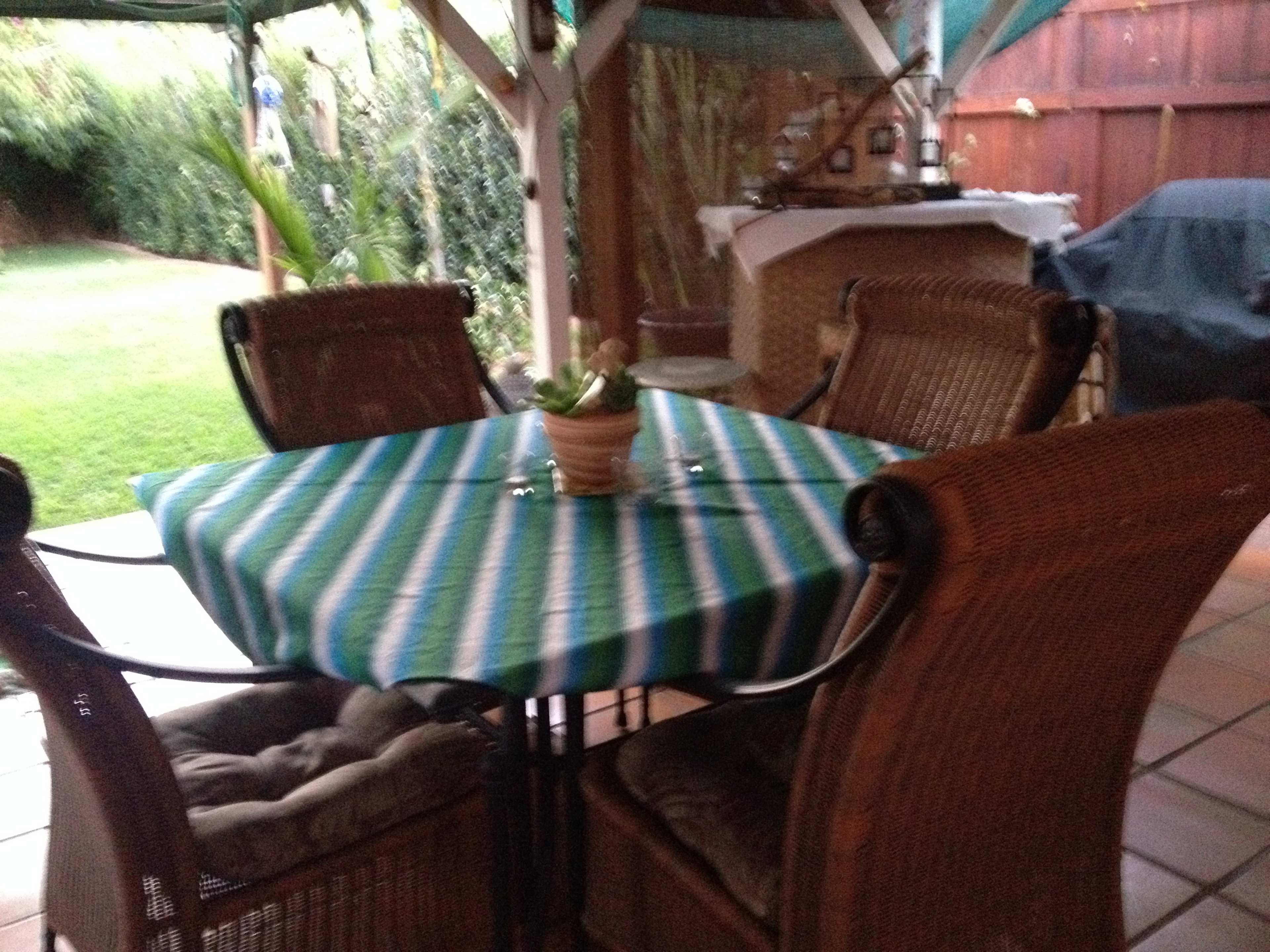 A patio table with a striped tablecloth is surrounded by four wicker chairs under a gazebo in a backyard.