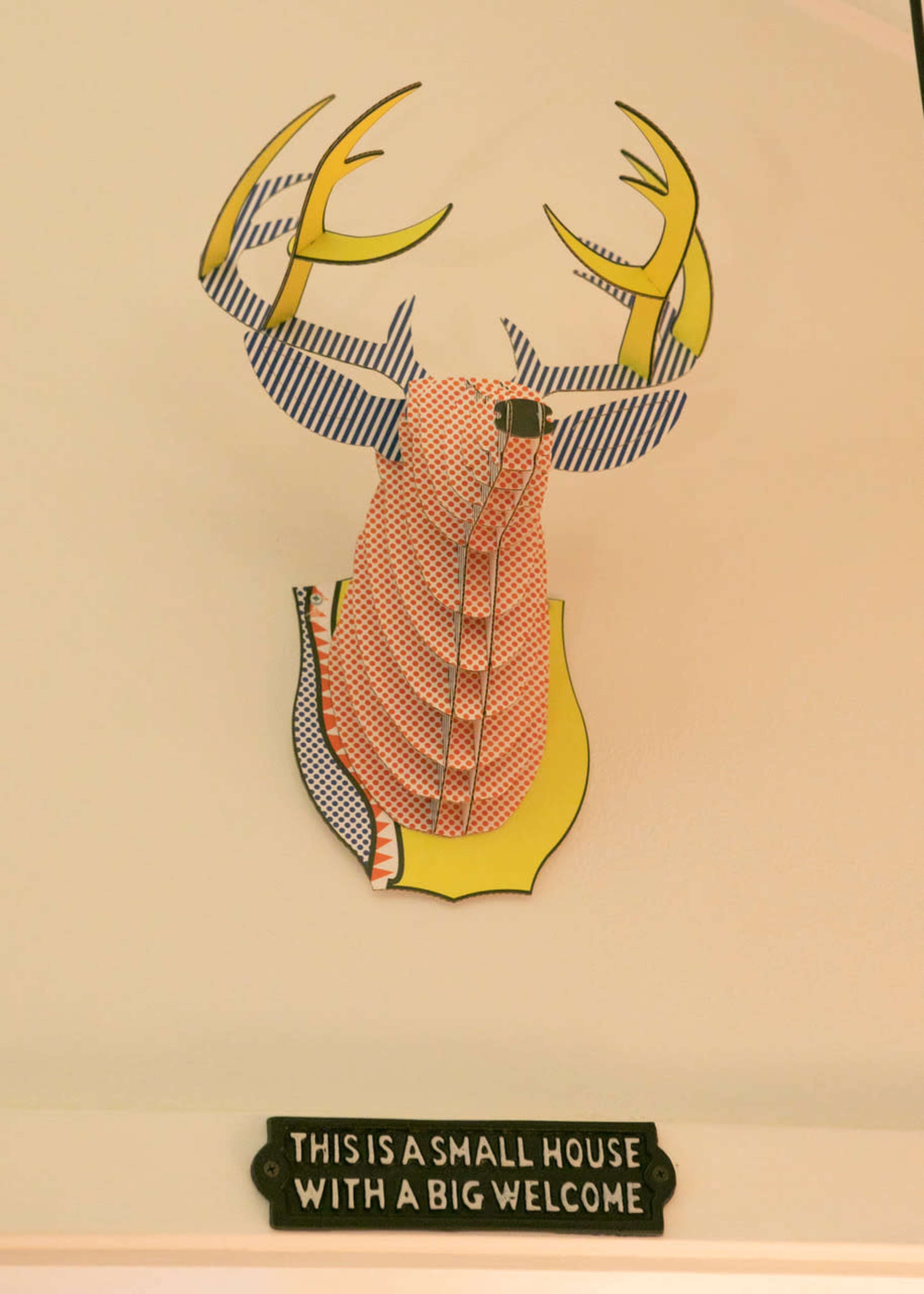 A colorful, artistic representation of a deer head mounted on a wall, adorned with a patterned shirt and accompanied by a sign that reads, "THIS IS A SMALL HOUSE WITH A BIG WELCOME."