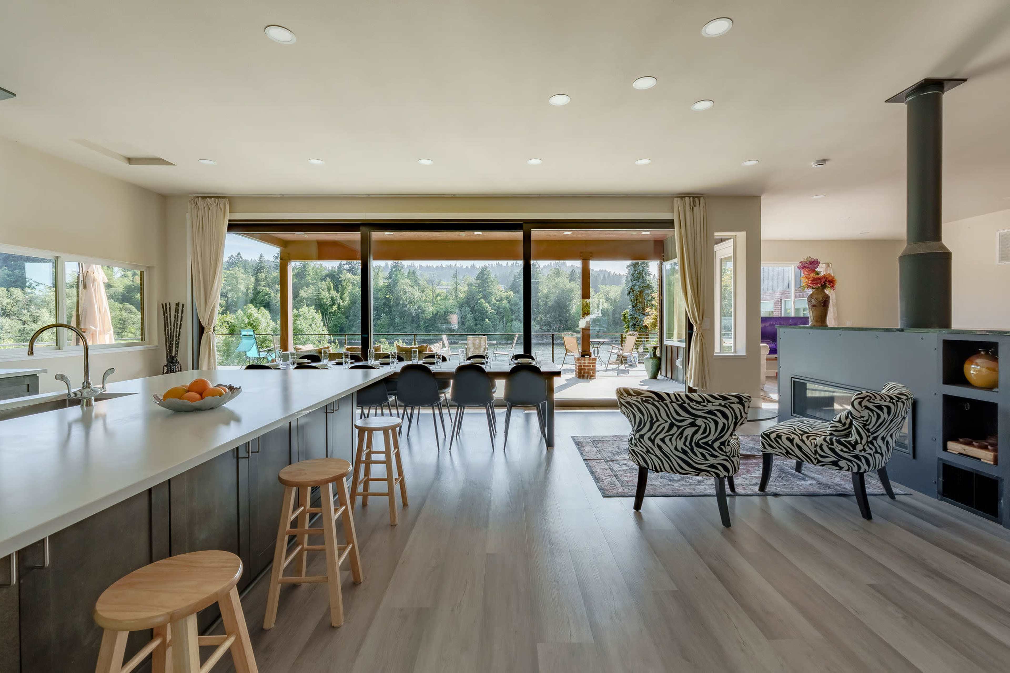 A modern kitchen with a large island, featuring bar stools, opens up to a spacious living area with large windows overlooking a green outdoor landscape.
