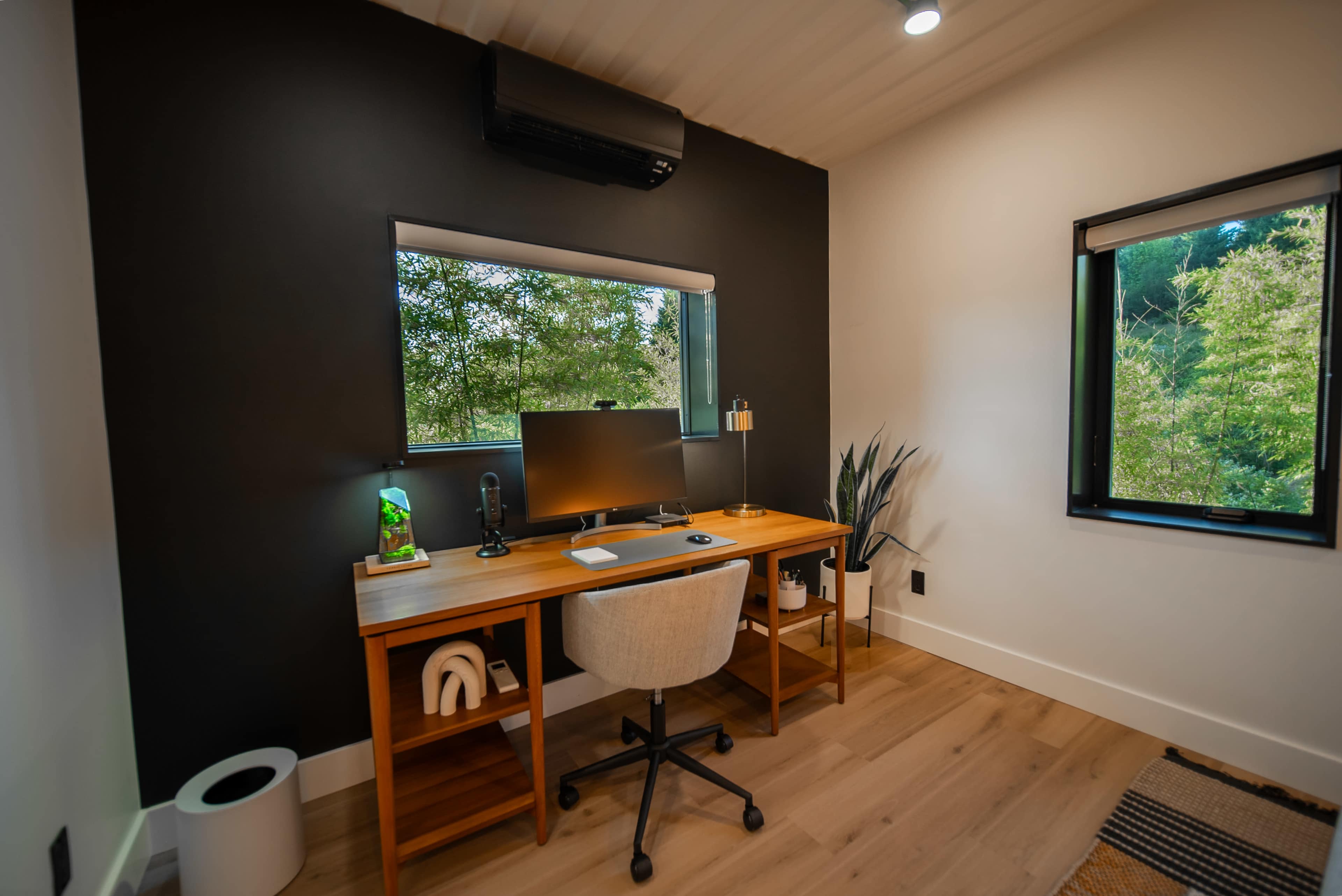 The image shows a modern home office with a wooden desk, a computer monitor, and a chair, situated next to a window overlooking greenery.