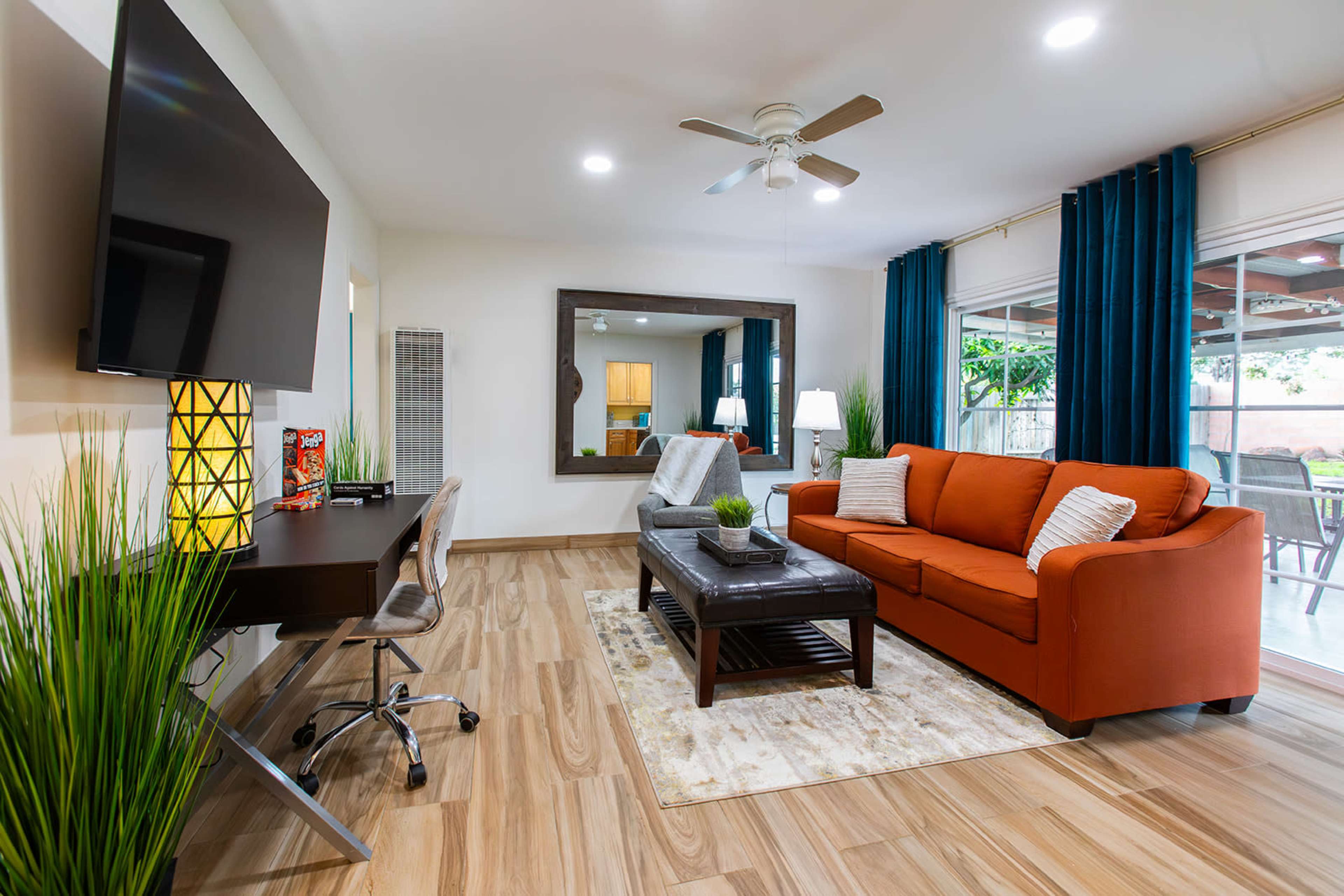 A modern living room featuring an orange sofa, a black coffee table, a desk with a chair, and large windows with blue curtains.