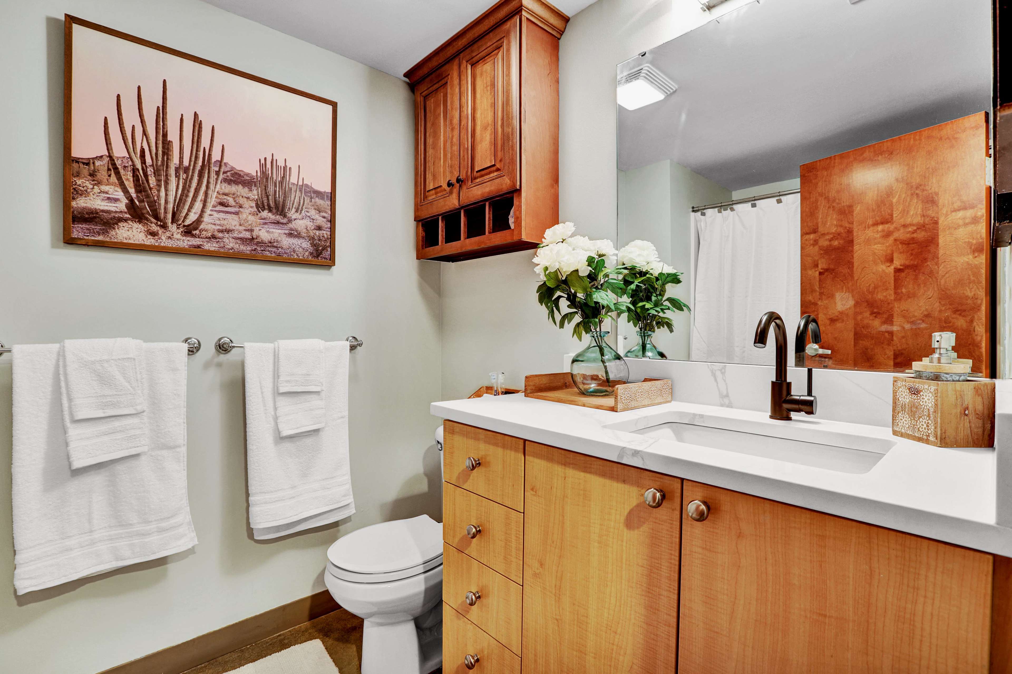The bathroom features a wooden cabinet, a white countertop sink, a framed cactus picture, and neatly hung towels.