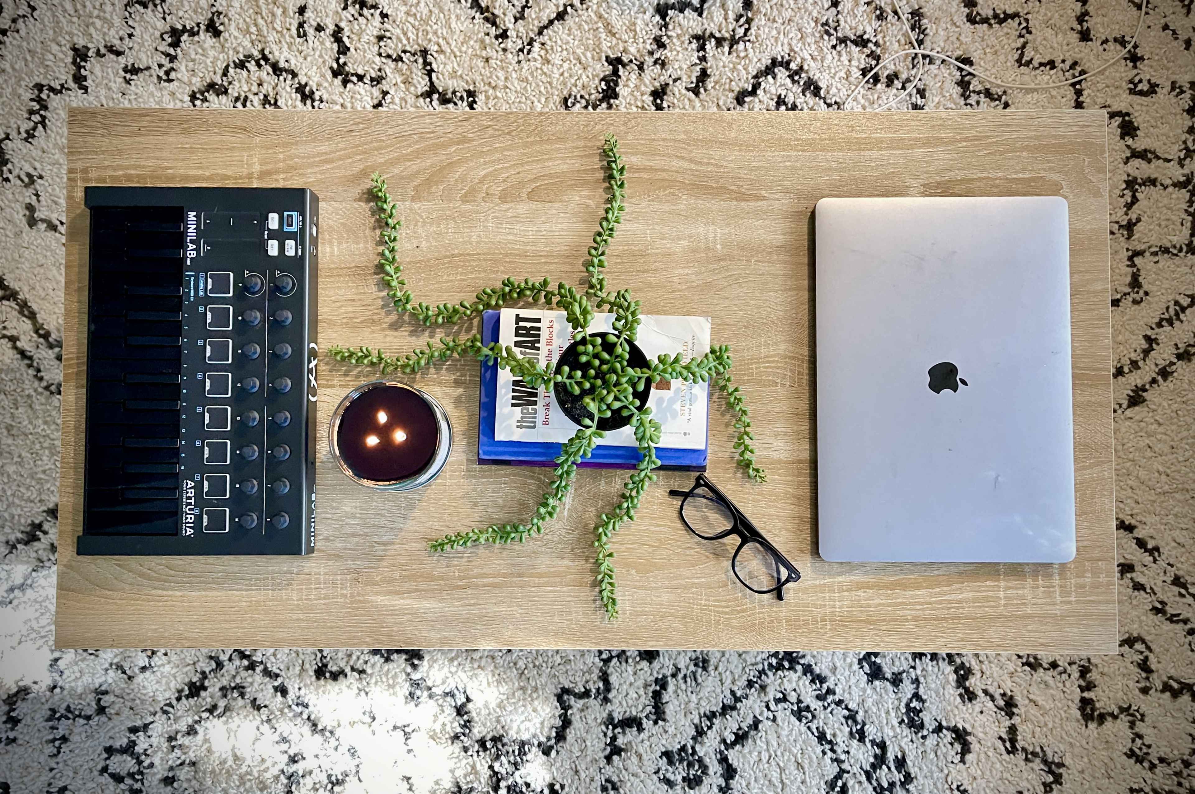 The image shows a wooden coffee table with a MIDI controller, a potted plant, a candle, a stack of books, a MacBook, and a pair of glasses arranged on its surface.