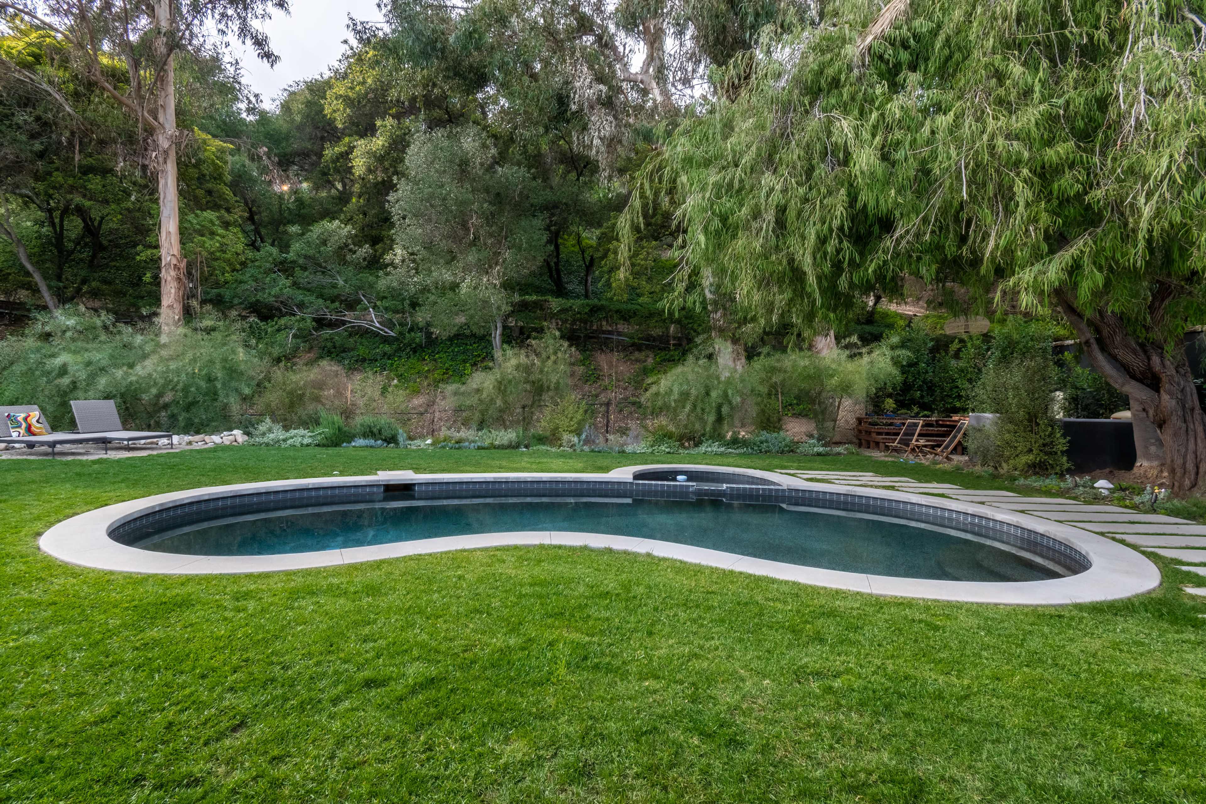 A kidney-shaped swimming pool is surrounded by green grass and lush foliage.