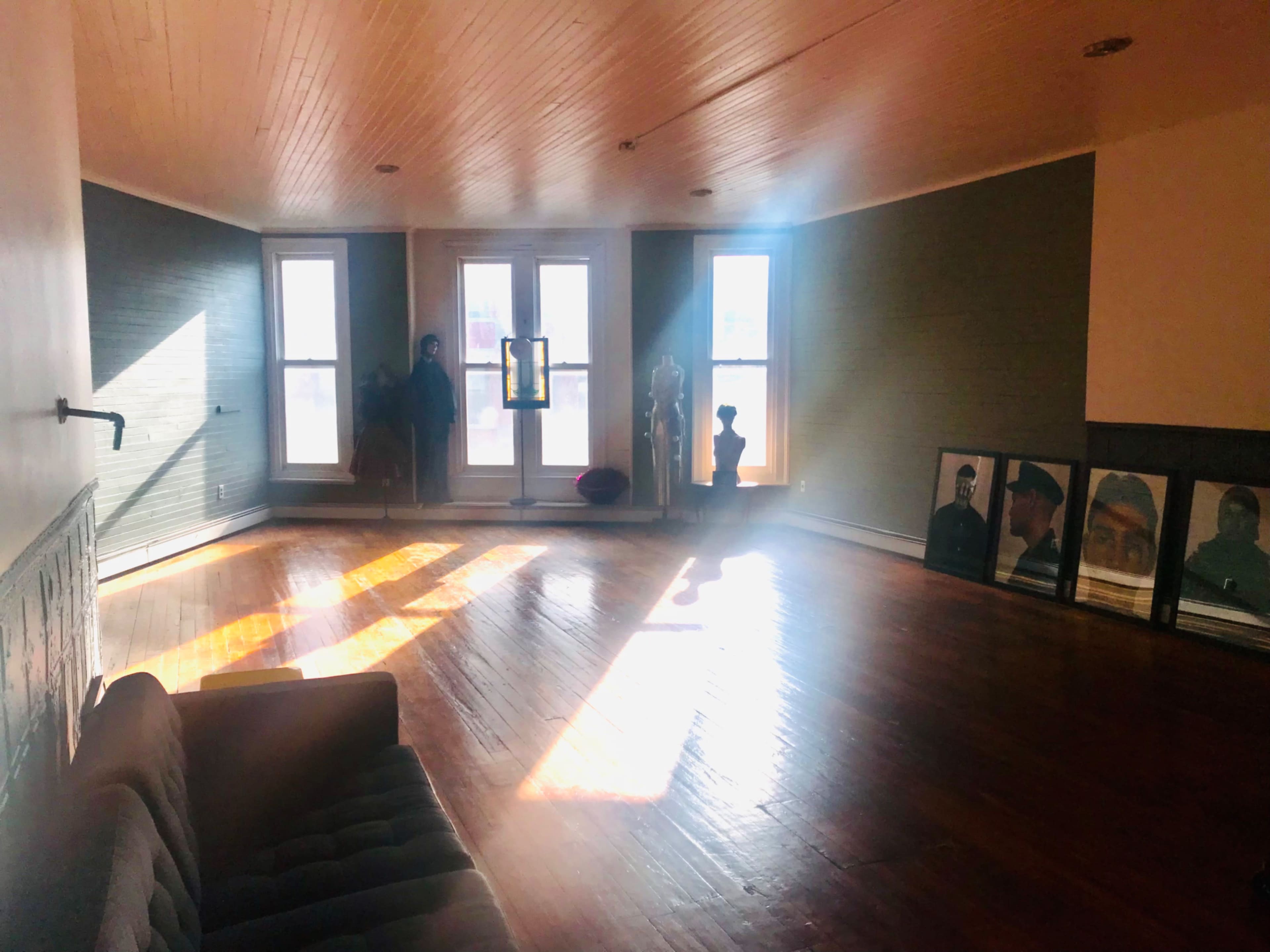 A spacious room with hardwood floors features large windows, a sofa, and framed pictures against the wall.