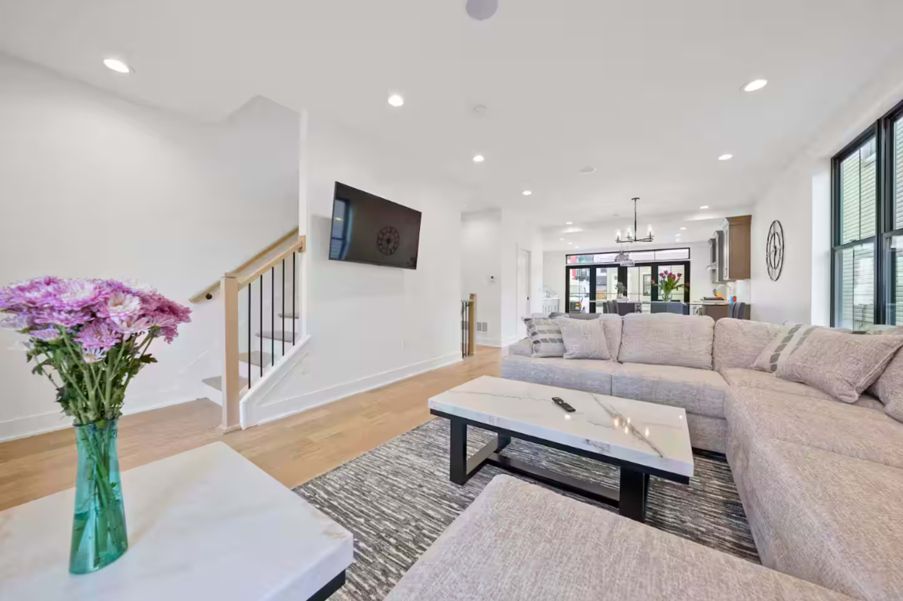 The living room features a large sectional sofa, a coffee table, and a staircase leading to an upper level, with a television mounted on the wall and natural light coming through large windows.