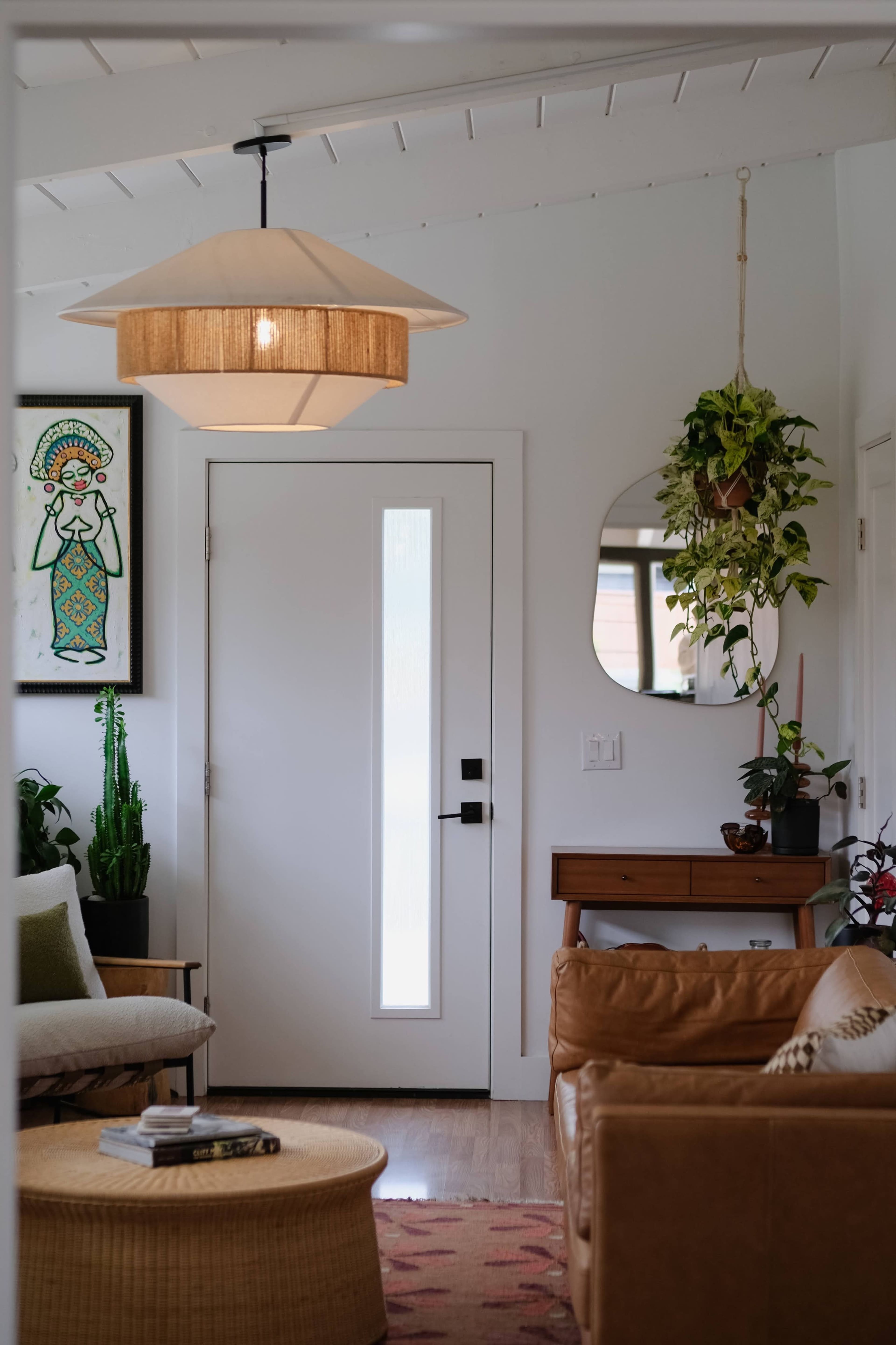 A well-lit living room features a stylish light fixture, a comfortable couch, and a door with a vertical window, complemented by plants and artwork on the walls.