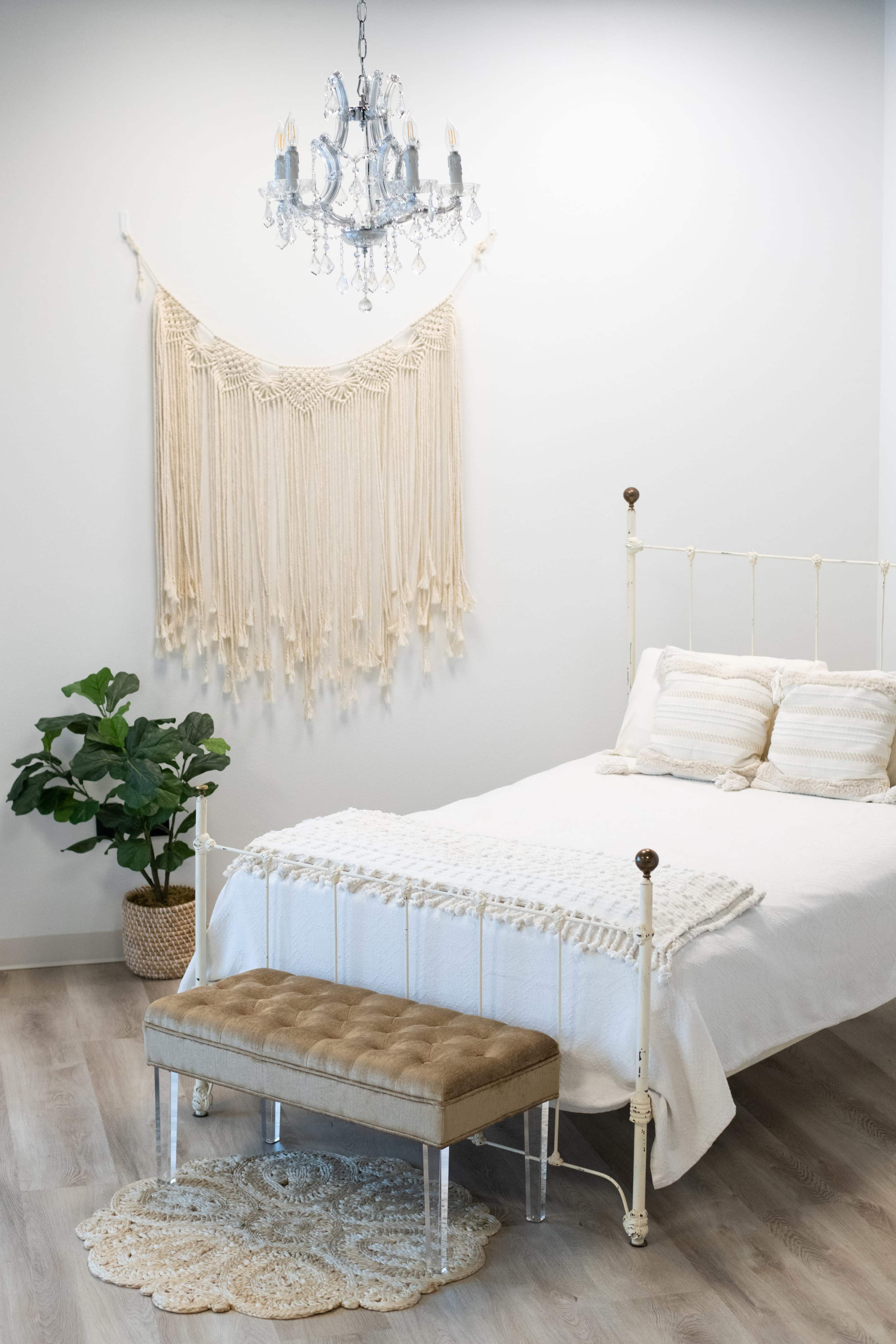 A neatly arranged bedroom features a white bed, a decorative macramé wall hanging, a crystal chandelier, and a plush bench at the foot of the bed.