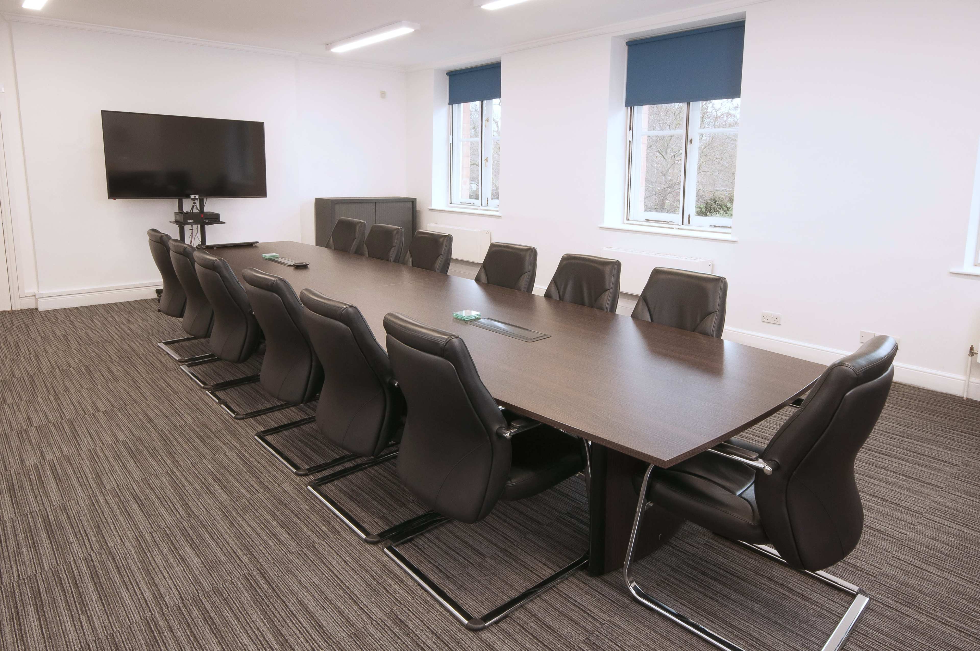 A long conference table with black leather chairs is set up in a bright, modern meeting room featuring a large wall-mounted screen and windows with blue shades.