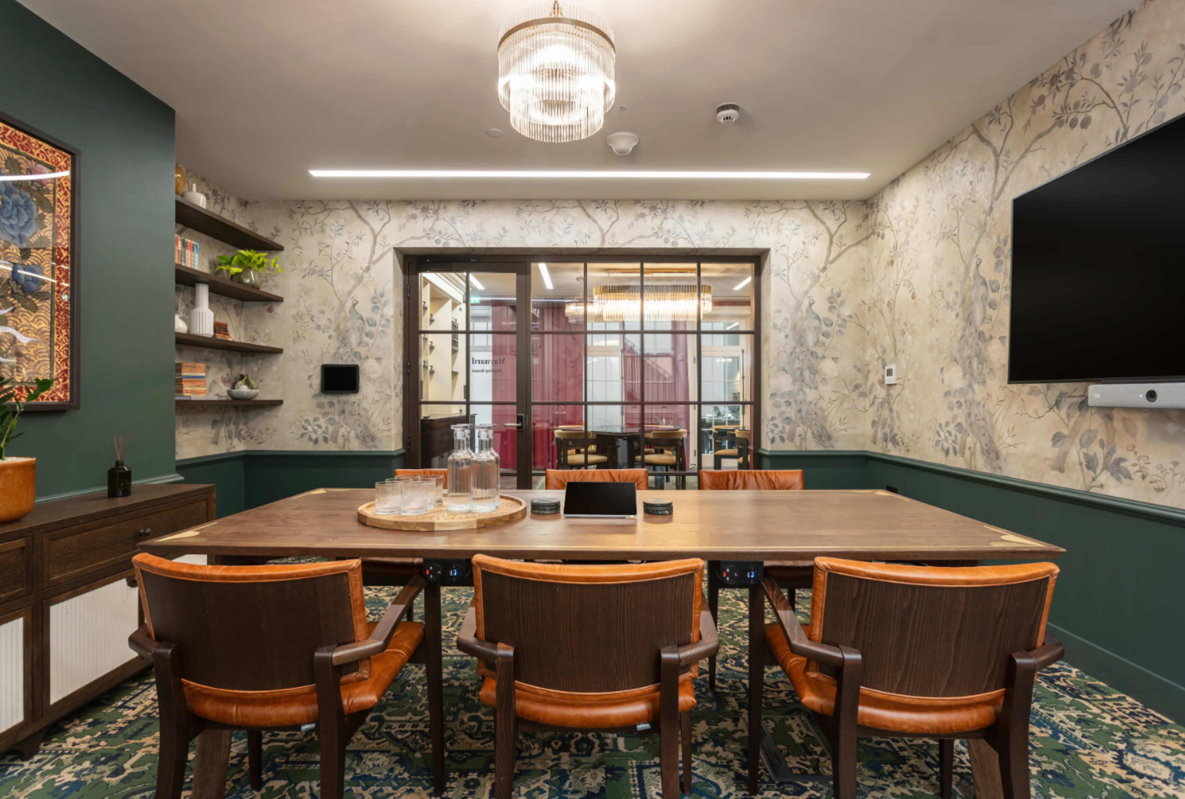 The image shows a conference room featuring a long wooden table surrounded by brown chairs, with floral wallpaper and a chandelier above.