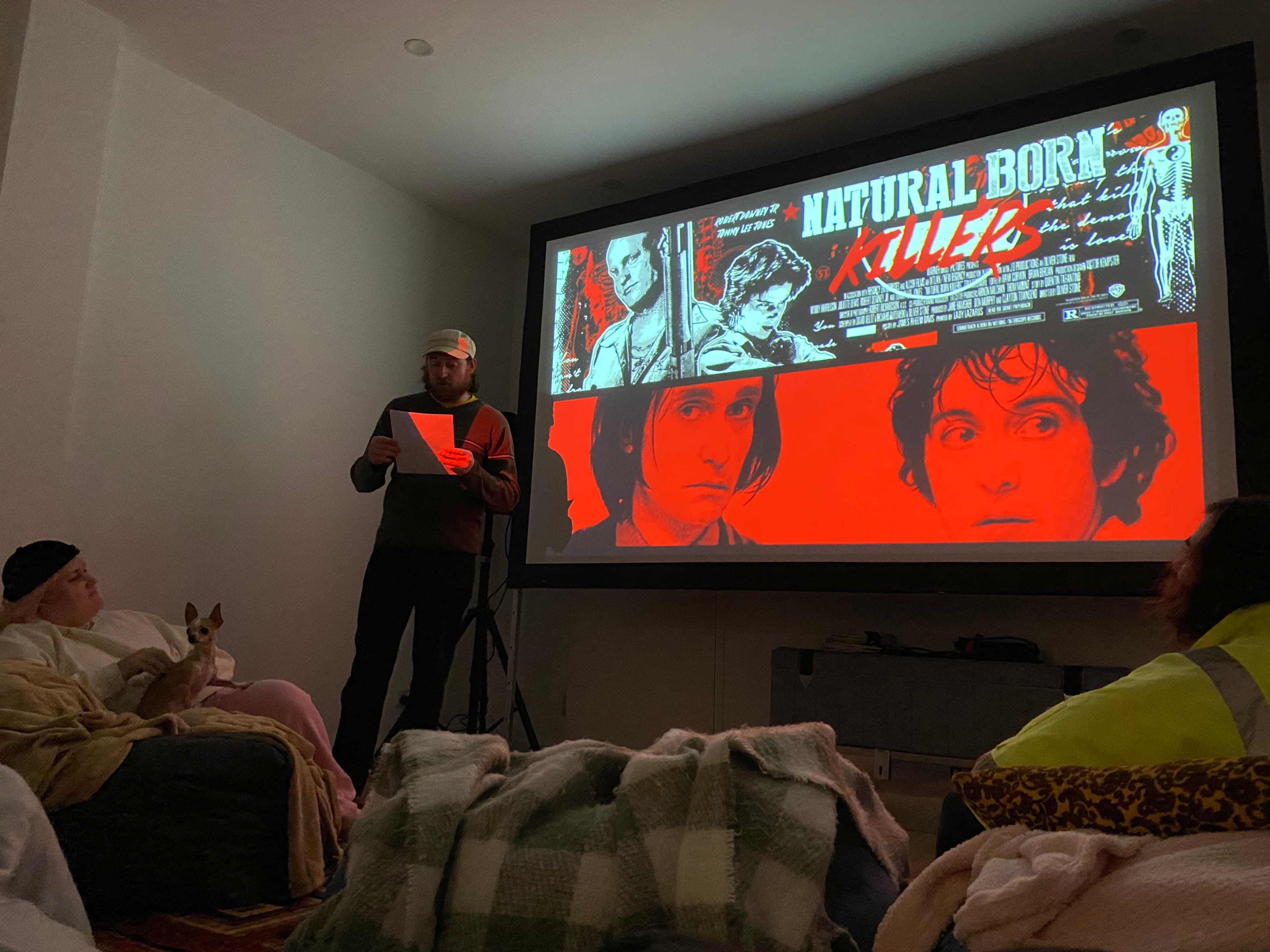A person stands reading from a paper in front of a large projected movie poster for "Natural Born Killers," while two others sit on a couch with a small dog.