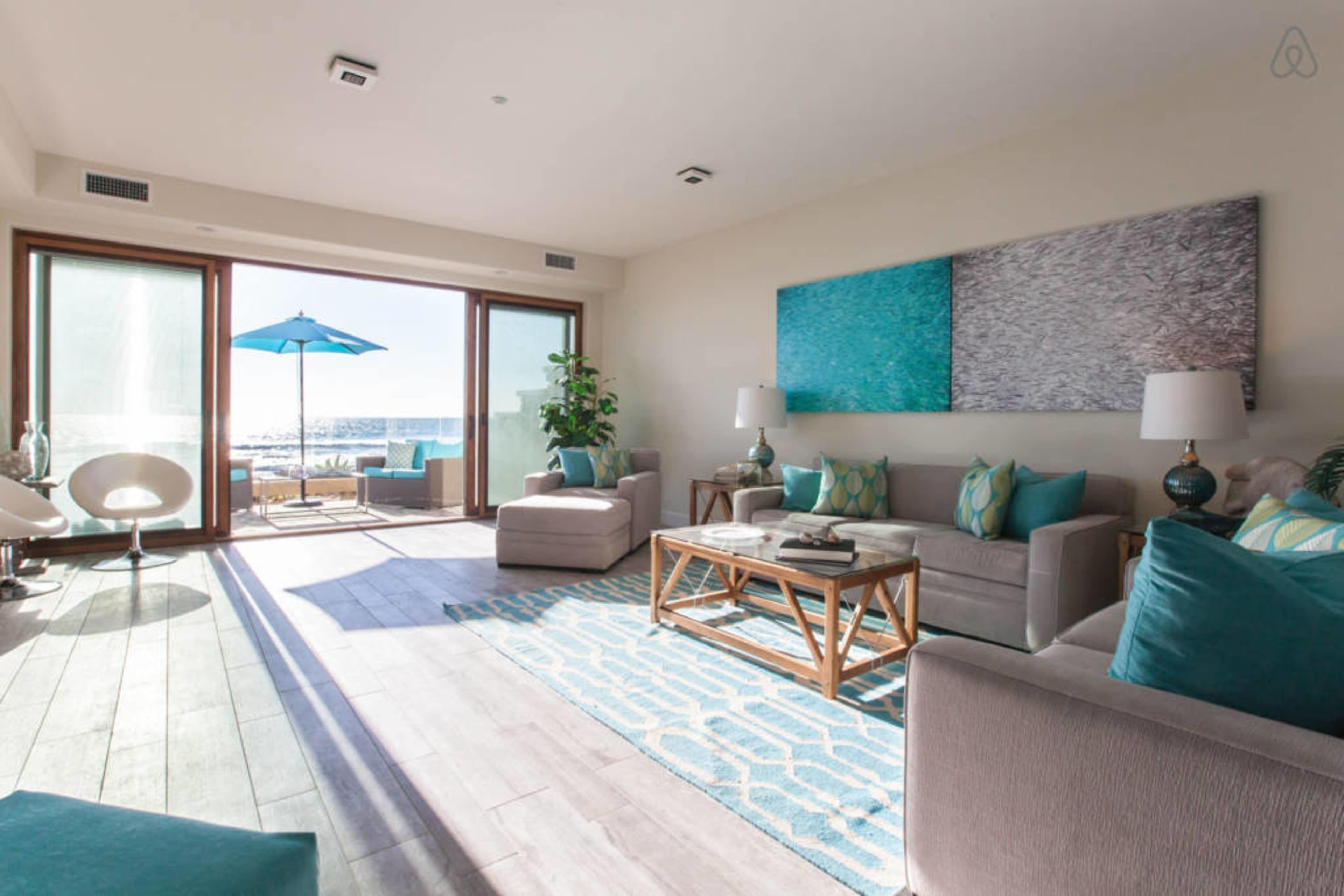 A modern living room with large windows overlooking the ocean, featuring two sofas, a coffee table, and an umbrella-covered outdoor seating area.