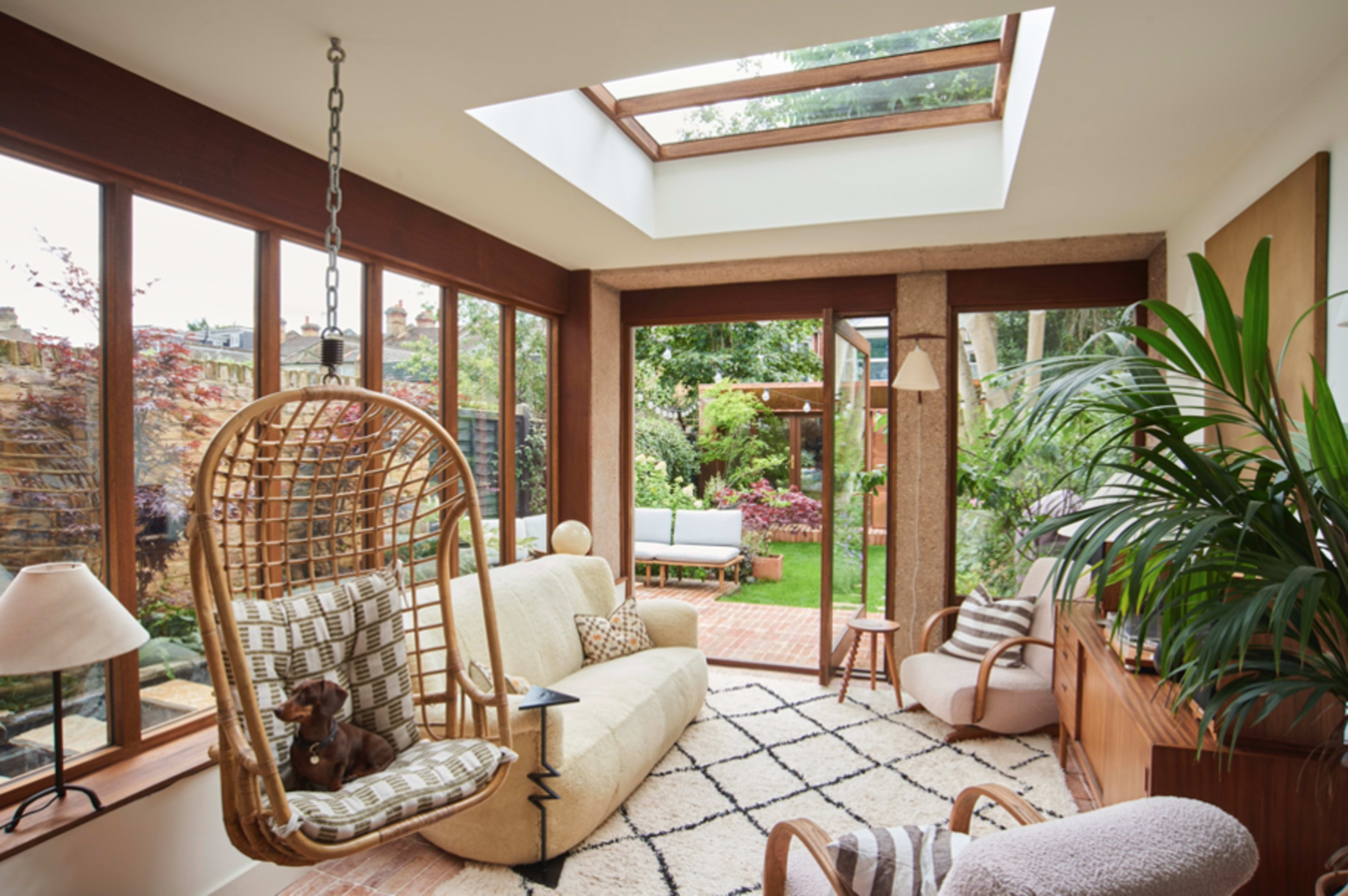 A sunlit living space features a hanging chair, a beige sofa, and large windows overlooking a garden.
