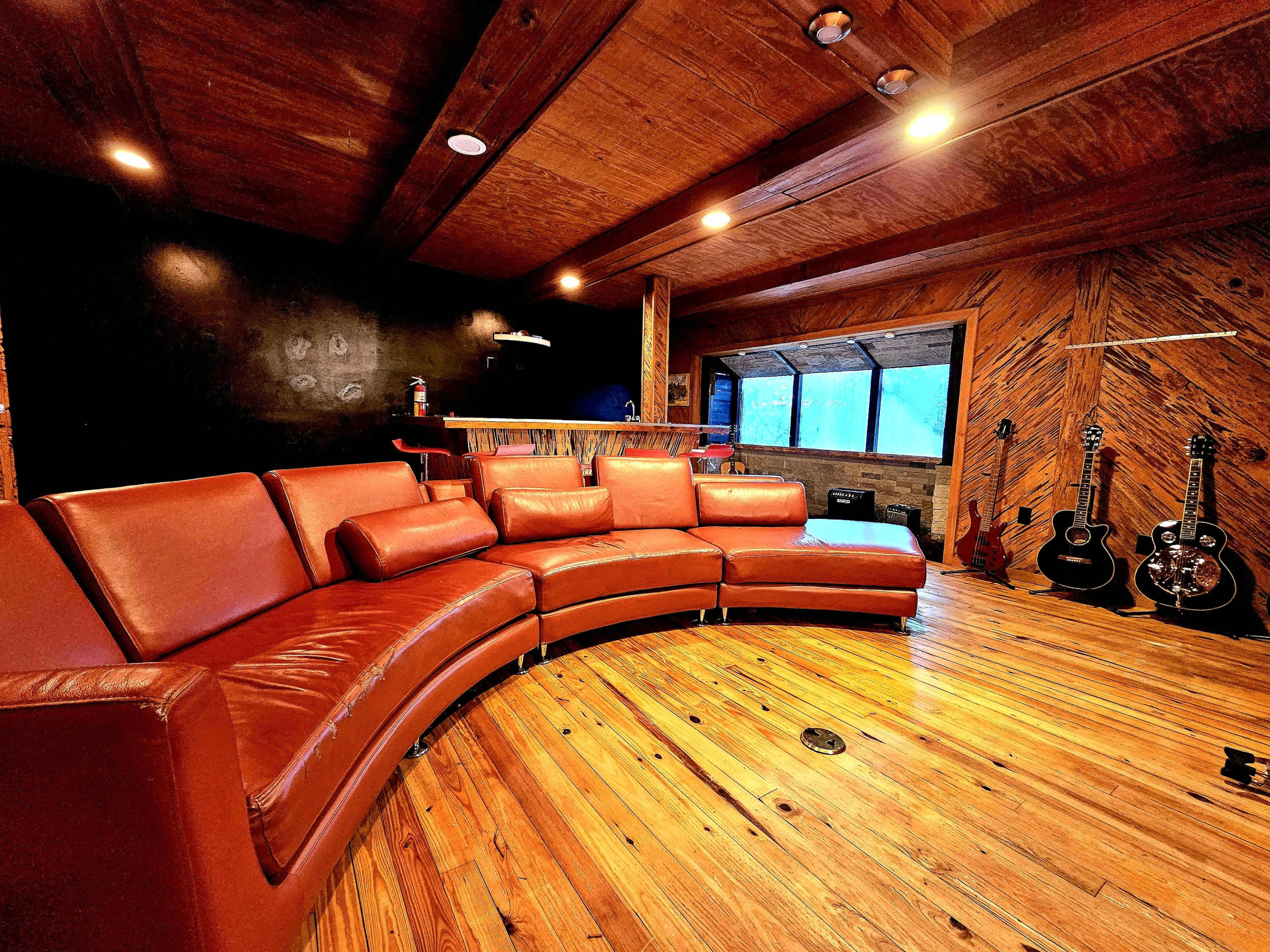 A curved brown leather sofa in a wooden-paneled room with guitars displayed on the wall and large windows letting in natural light.