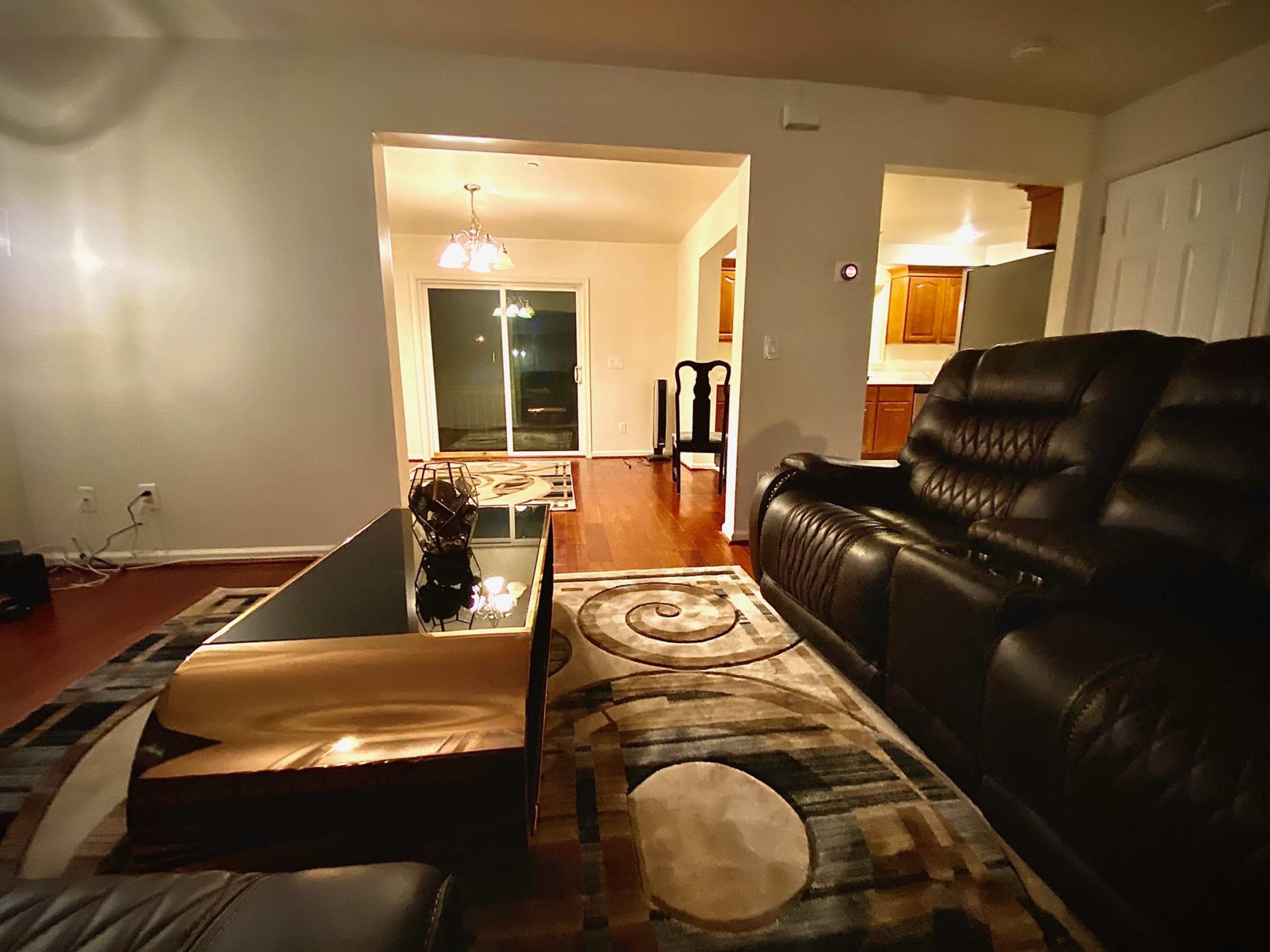 A living room with black leather seating, a coffee table, and a view into a space with hardwood floors and a sliding glass door.