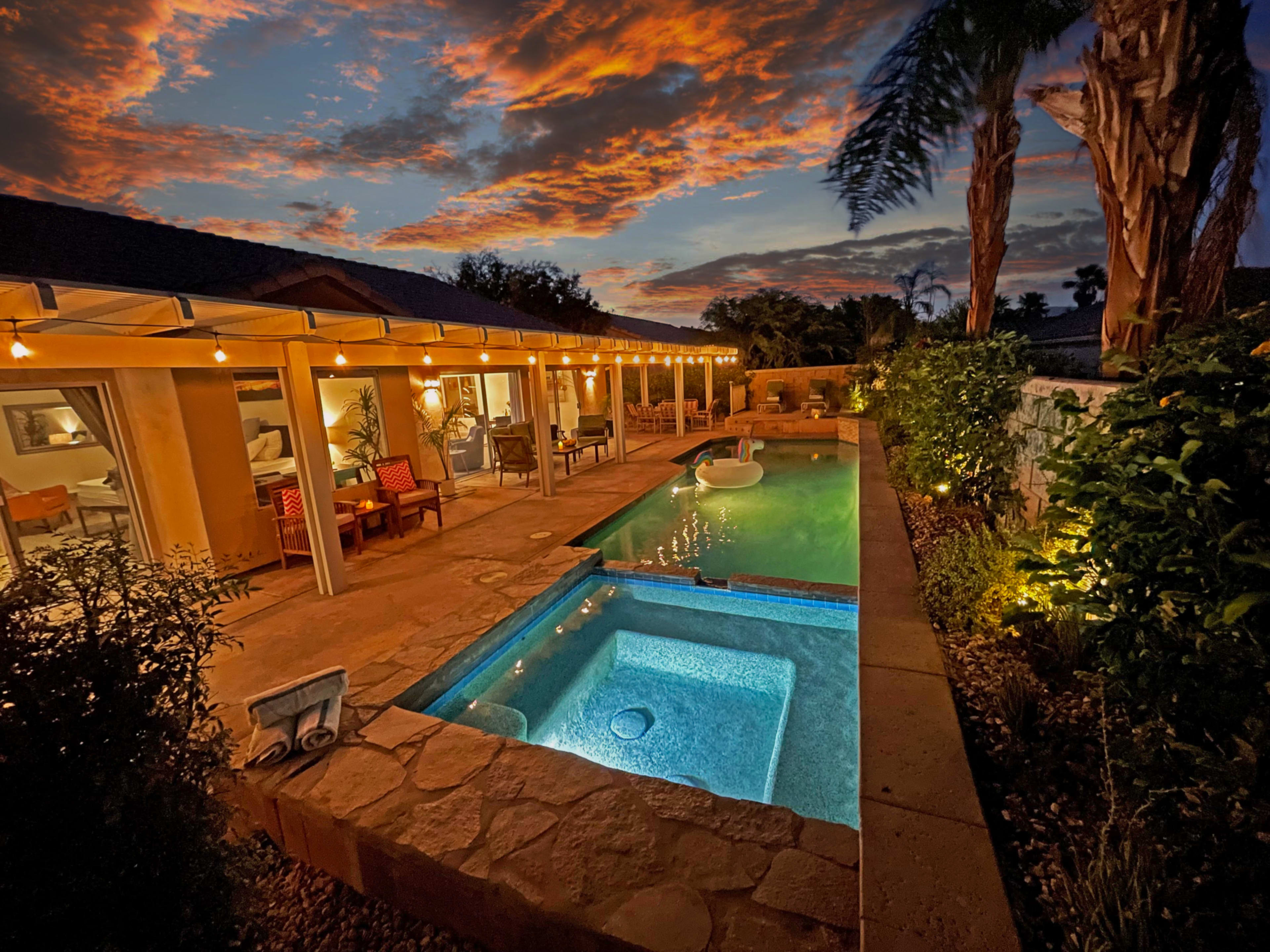 A well-lit patio with a swimming pool and spa is set against a vibrant sunset sky.