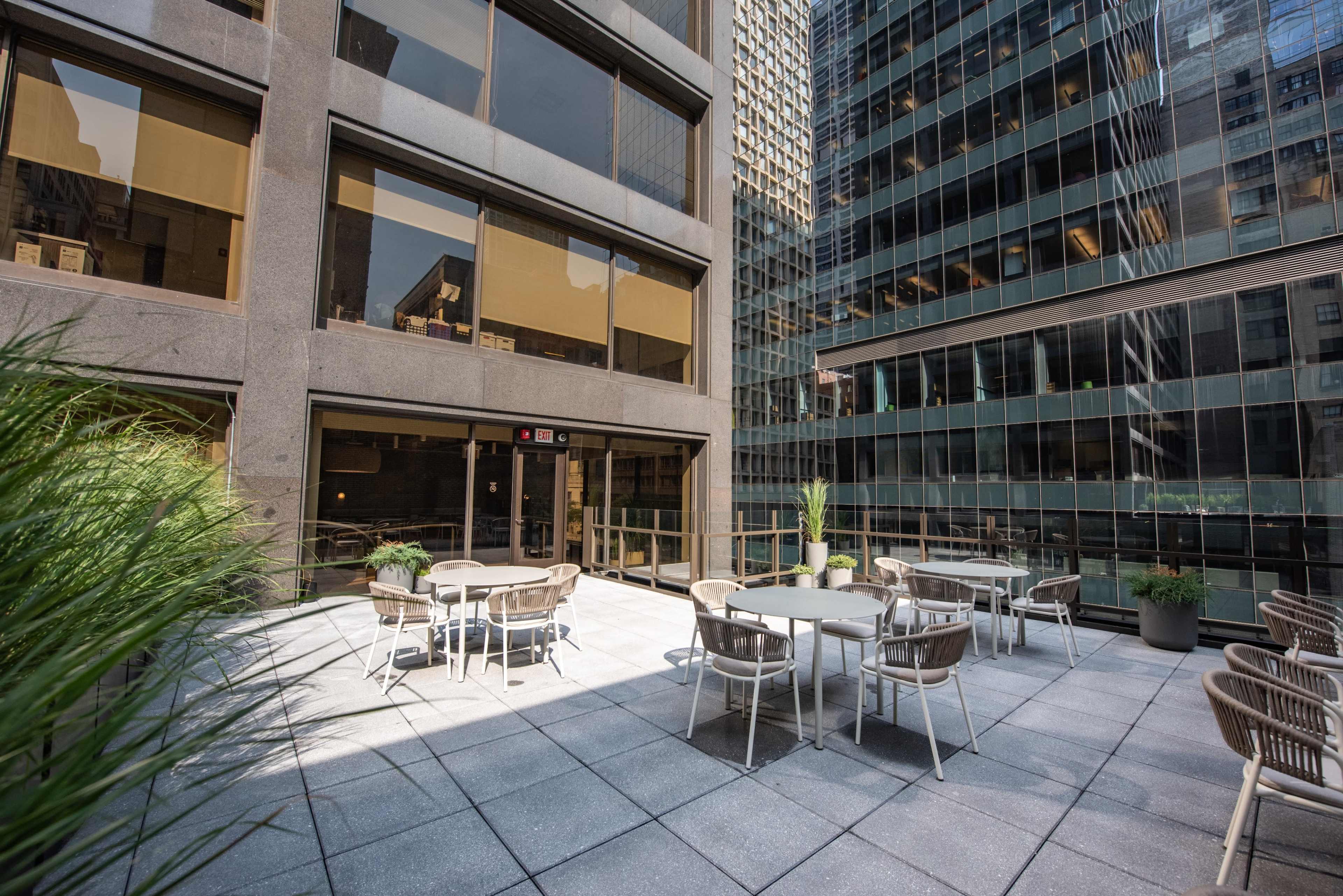 A concrete outdoor terrace features tables and chairs surrounded by tall office buildings.