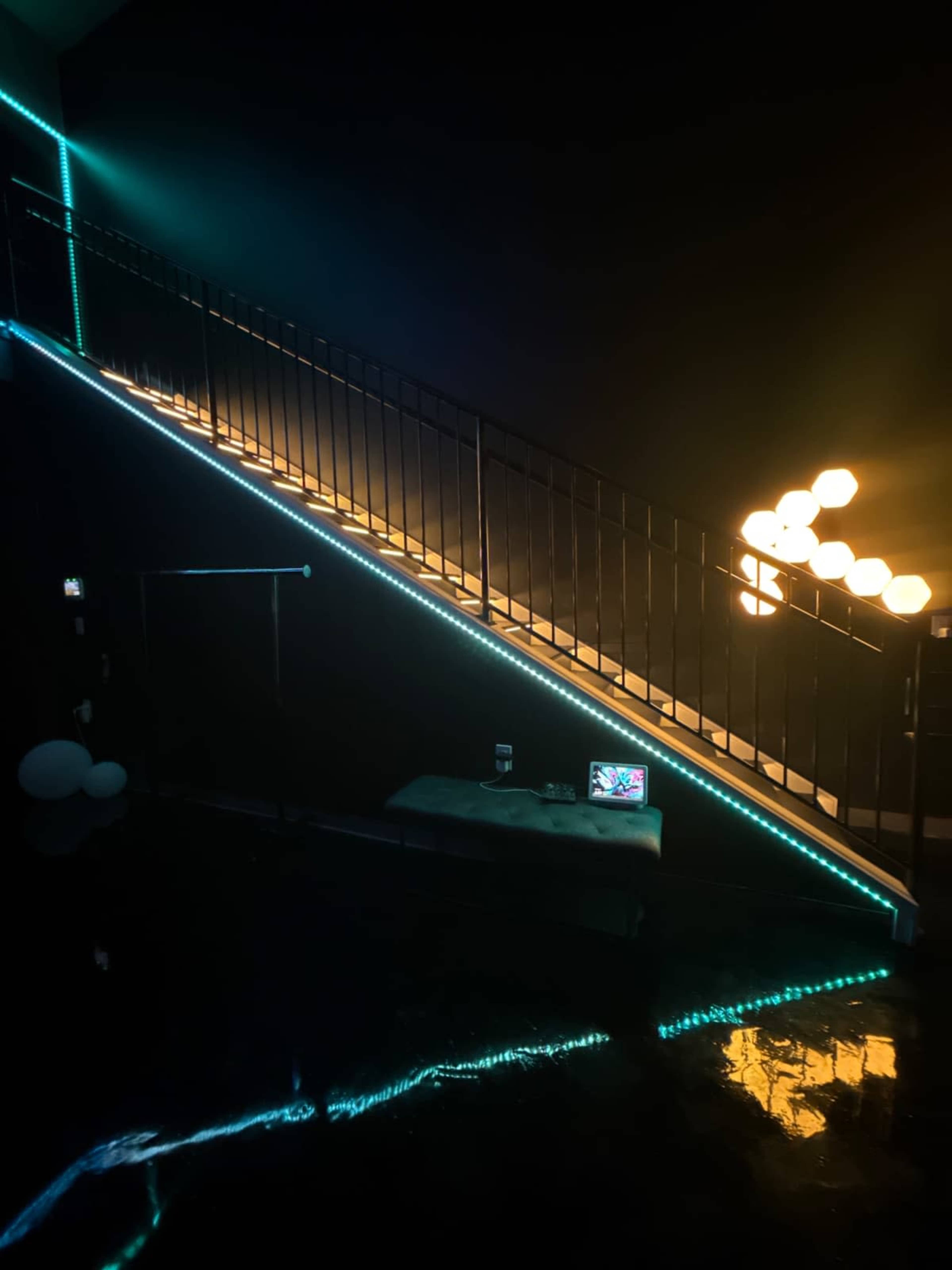 A staircase illuminated with colored LEDs ascends against a dark wall, accompanied by a table with a laptop and decorative lights in the background.