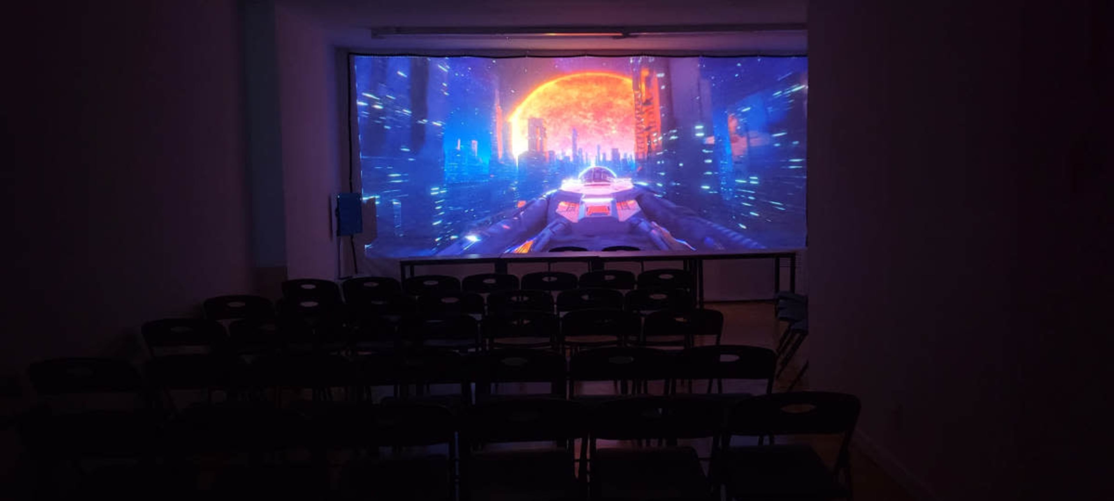 The image shows a dark room with rows of empty black chairs facing a large screen displaying a colorful futuristic cityscape with a glowing orange sun and blue lights.