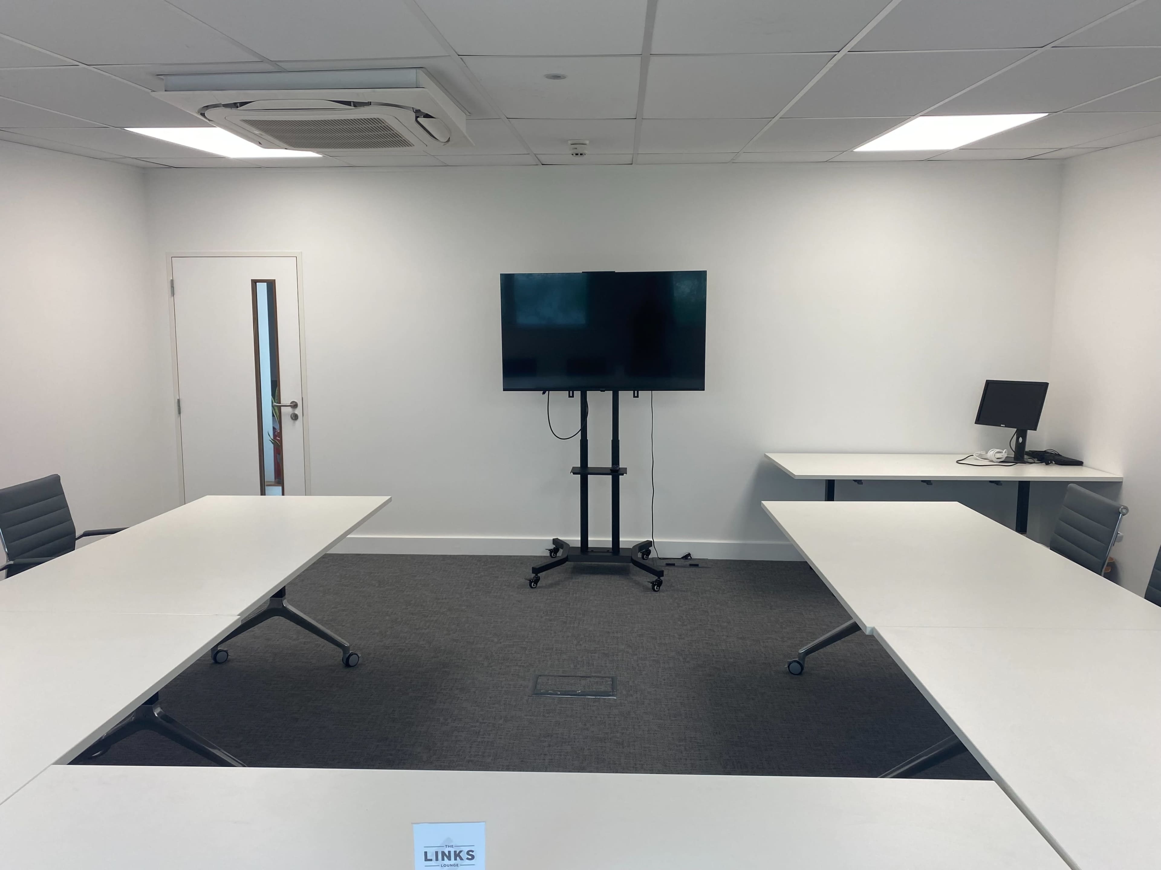 A video conference room with two tables arranged in an L-shape, a wall-mounted TV, and a computer workstation.